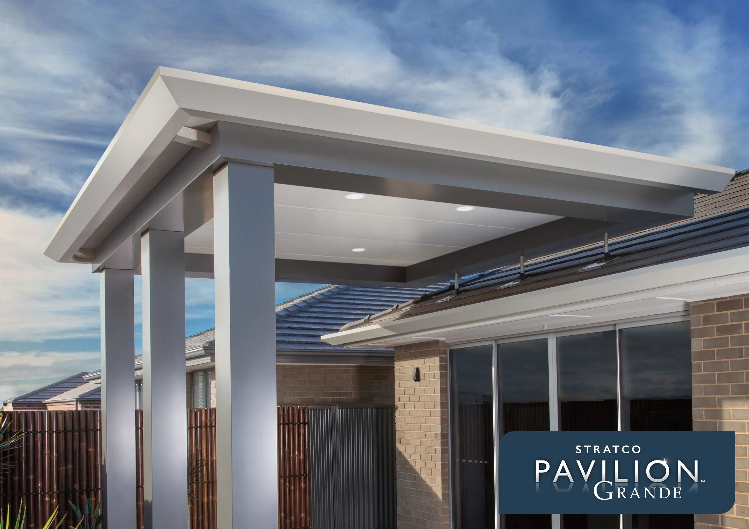 Exterior view of a modern house with a white patio roof extending from the roof, with a blue sky and clouds in the background. The house has brick walls, large windows, and a fence. Platinum Decks authorised installer.