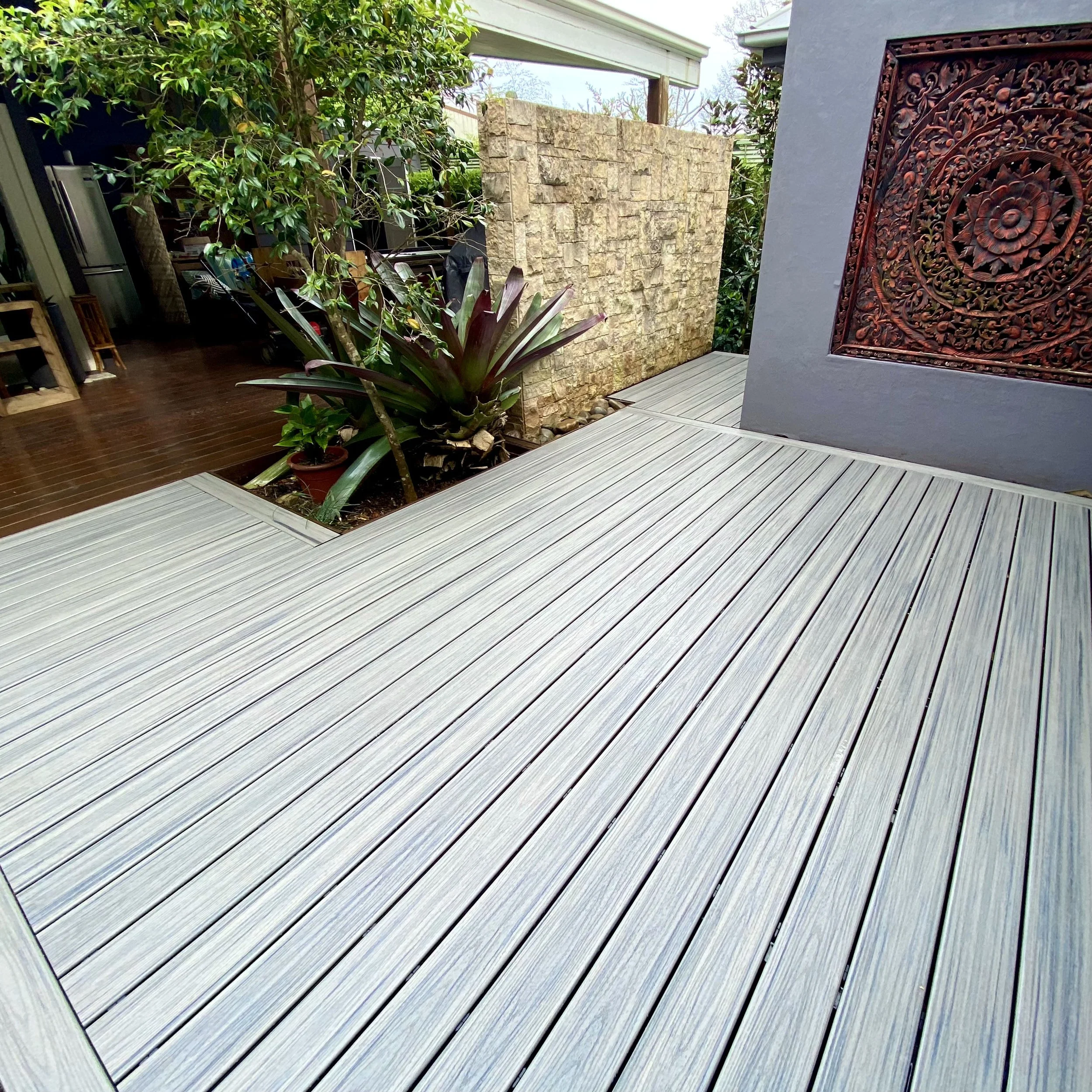 Outdoor patio area with wooden decking, potted plants including a large-leafed plant, a stone wall, and a decorative carved wooden panel on the wall.