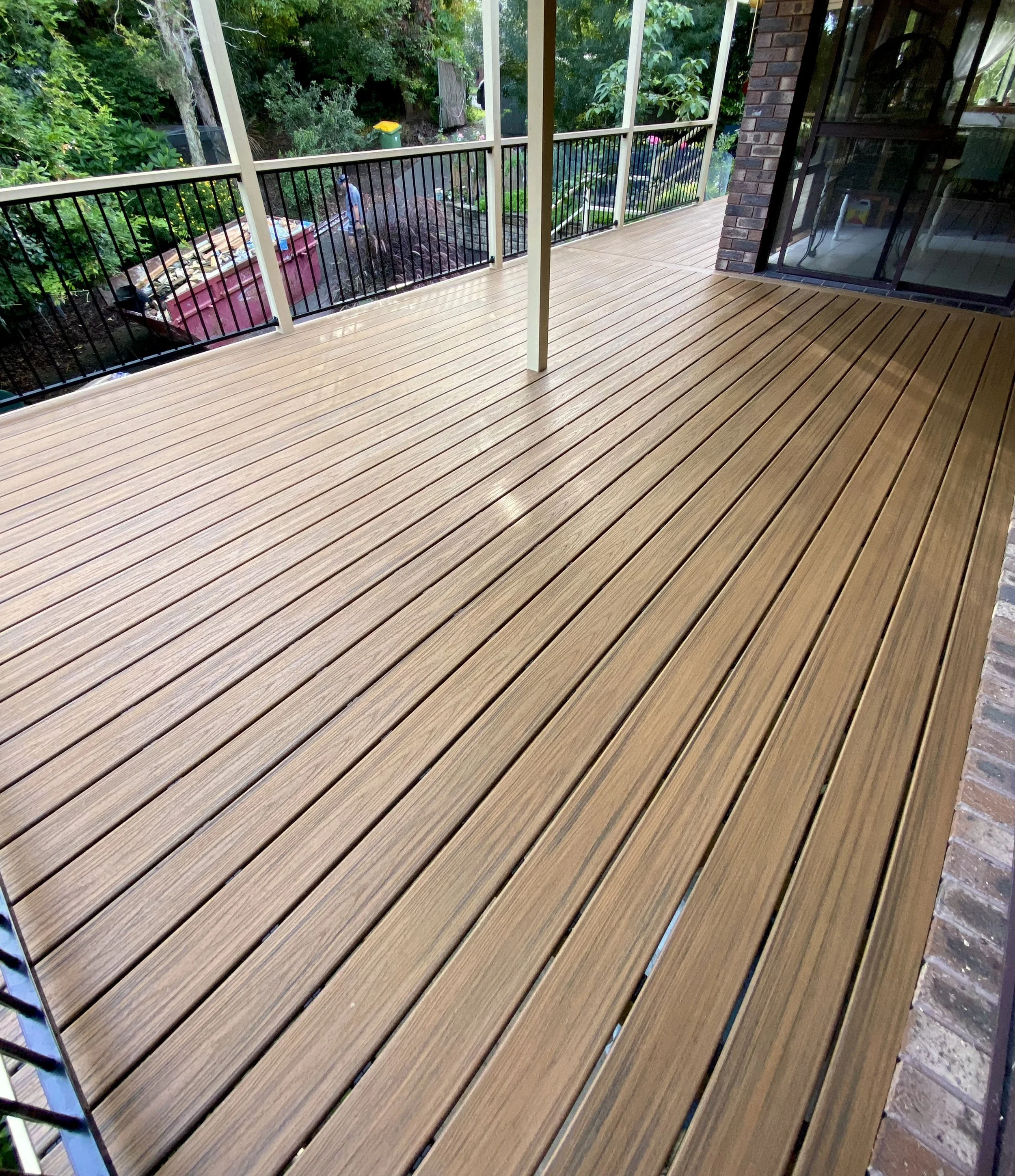 View from a porch or balcony with wooden flooring, black metal railing, and a screened enclosure, overlooking a backyard with trees, plants, and a person walking near a garden bed or compost area.