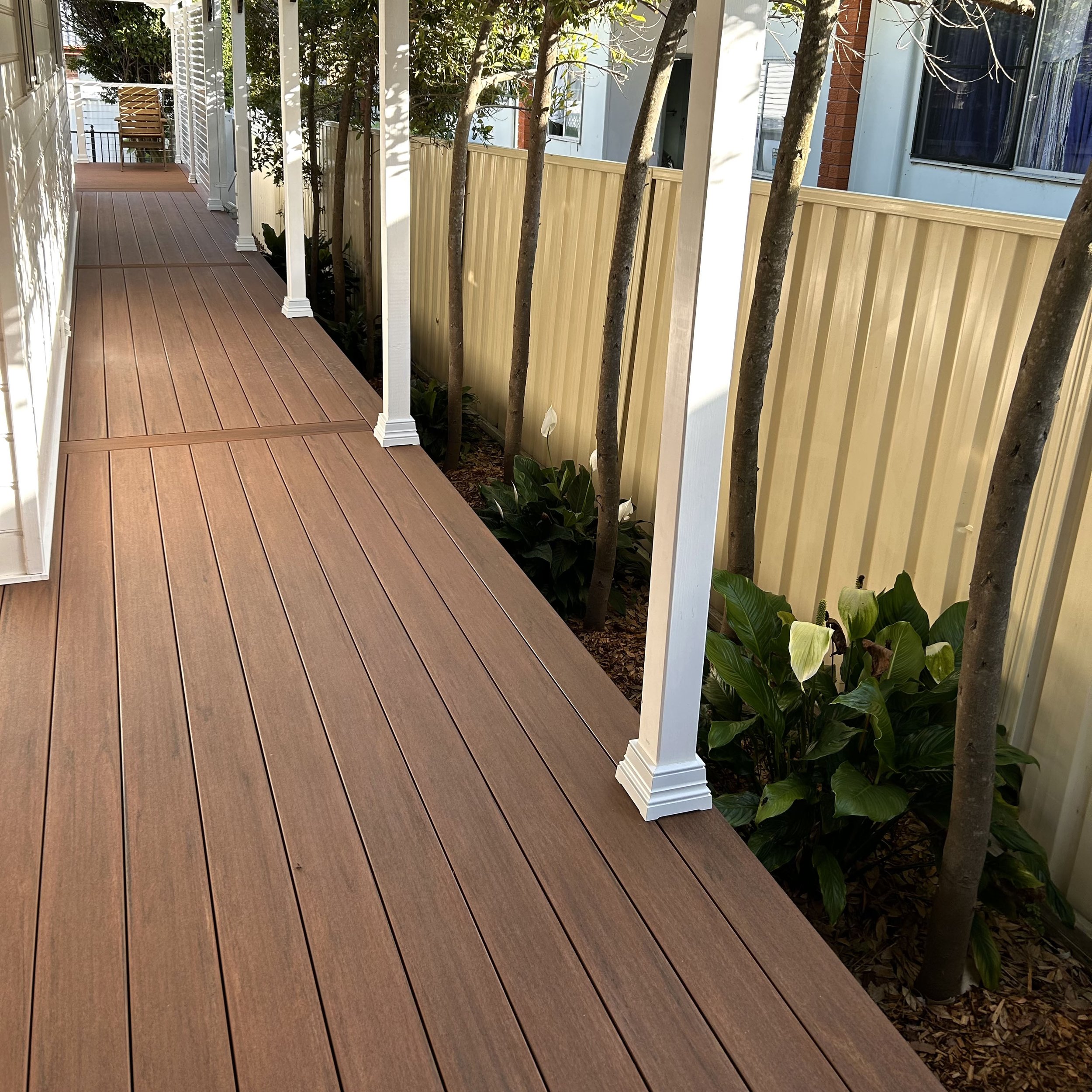 View of a composite outdoor deck with a white railing, trees, plants, and a yellow privacy fence along the side of a house with a window and brick wall.