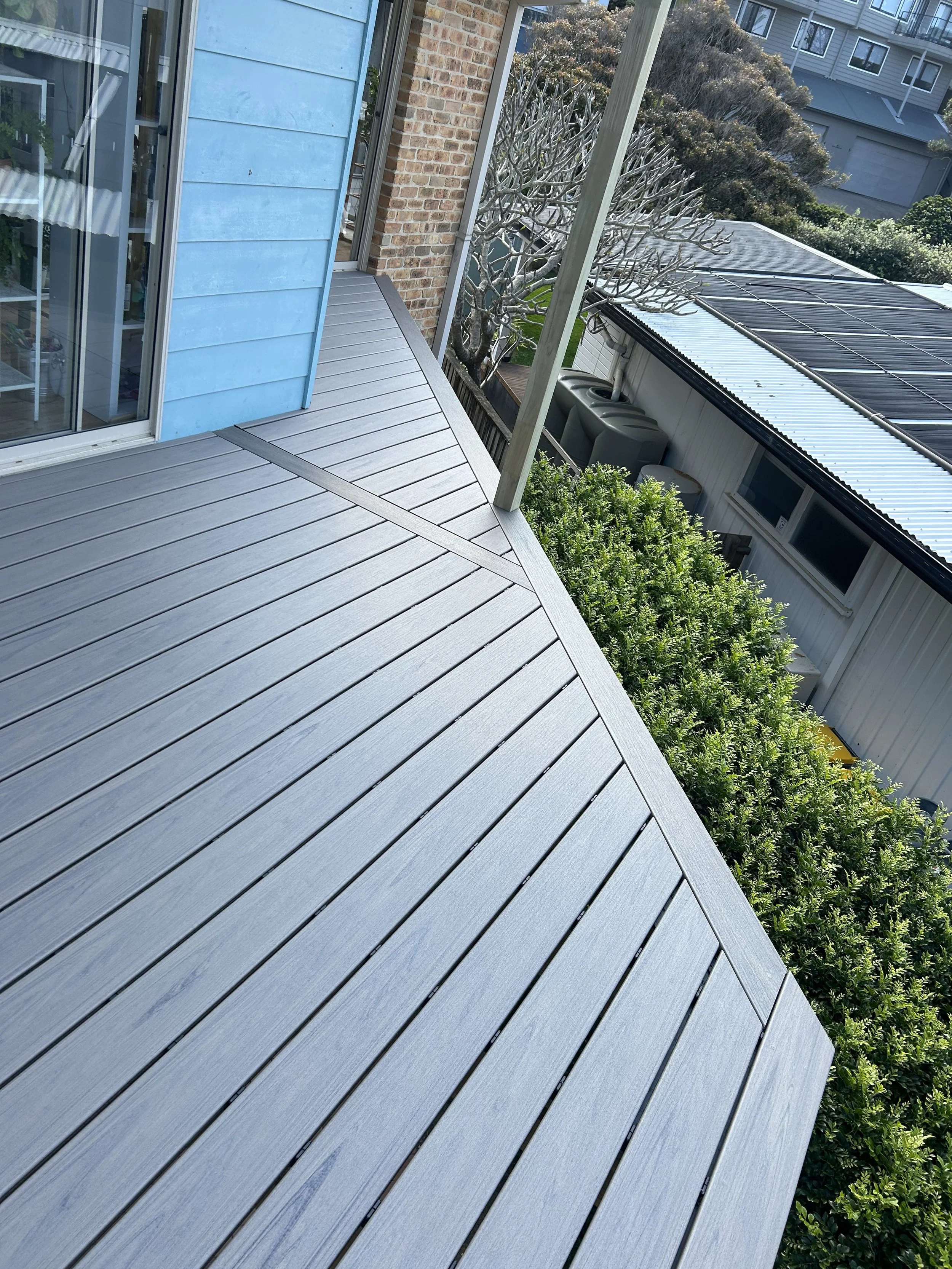 A backyard deck made of light gray composite wood with a glass sliding door leading into a house. There are neighboring houses and a garden with bushes and a tree with no leaves.