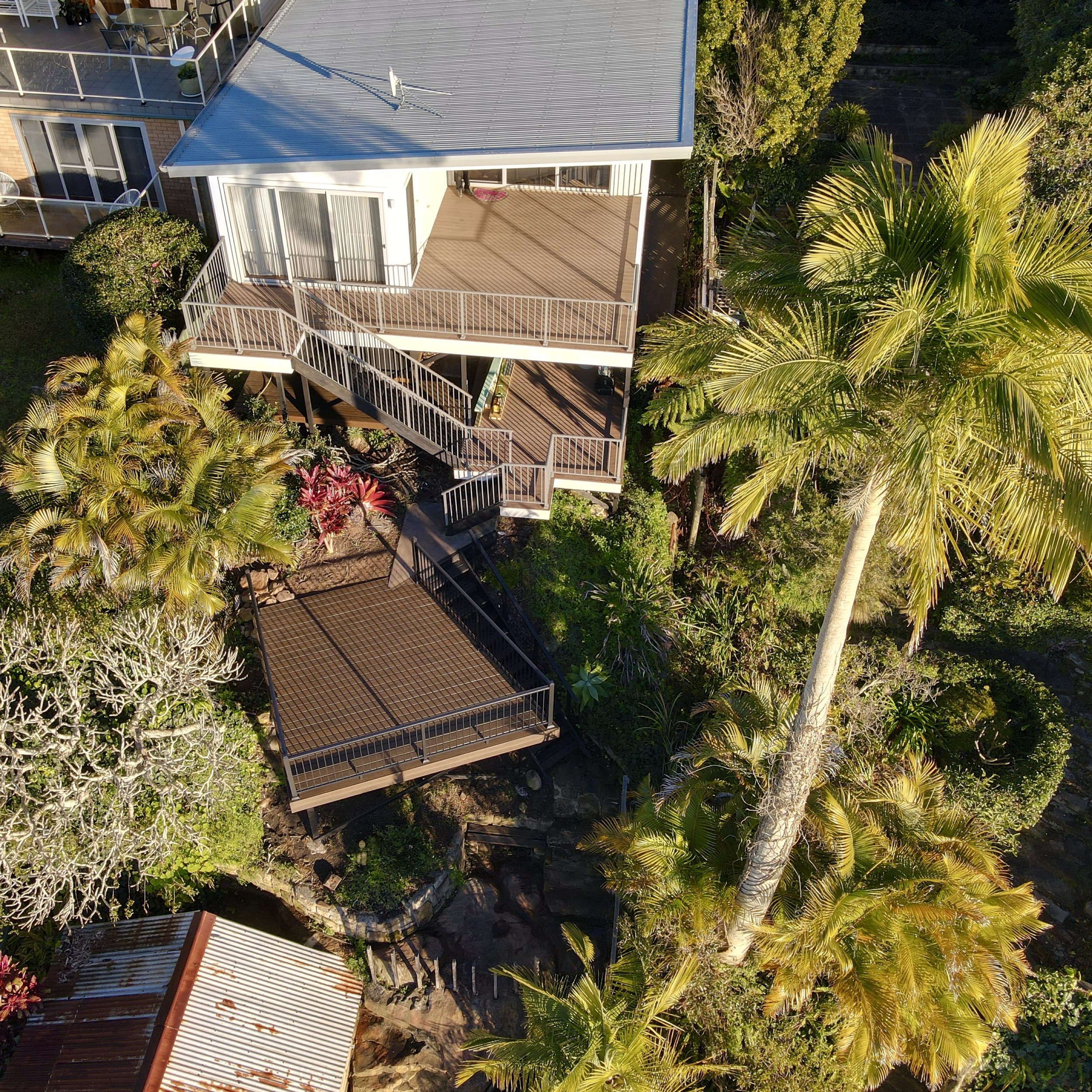An aerial view of a multi-level house surrounded by lush green tropical trees and plants, with a metal roof, Trex deck in spiced rum, and outdoor seating area.