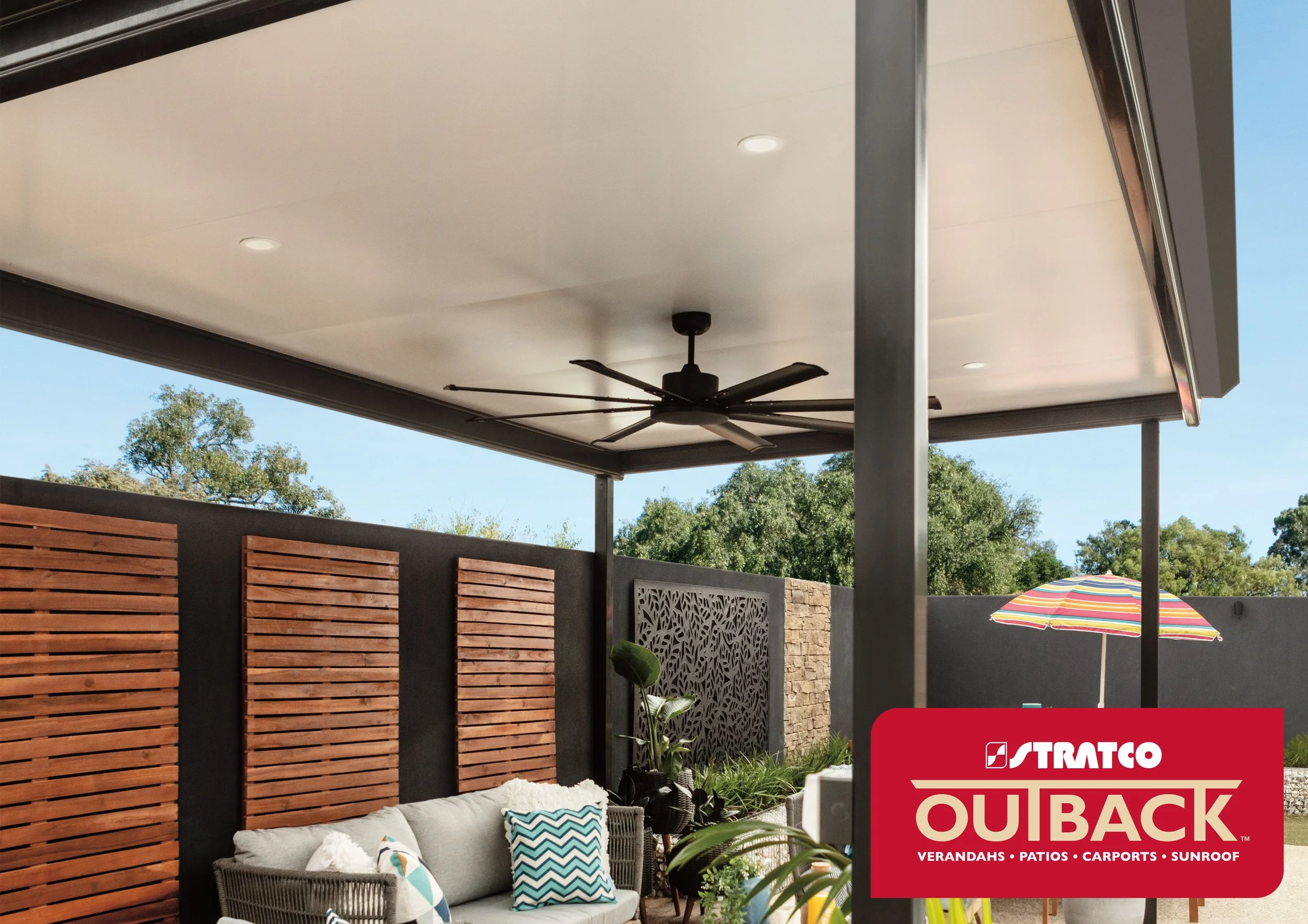Outdoor patio with a ceiling fan, wooden privacy screens, a colourful striped umbrella, and a seating area with pillows, under a covered verandah. Platinum Decks authorised Stratco Outback Dealer.