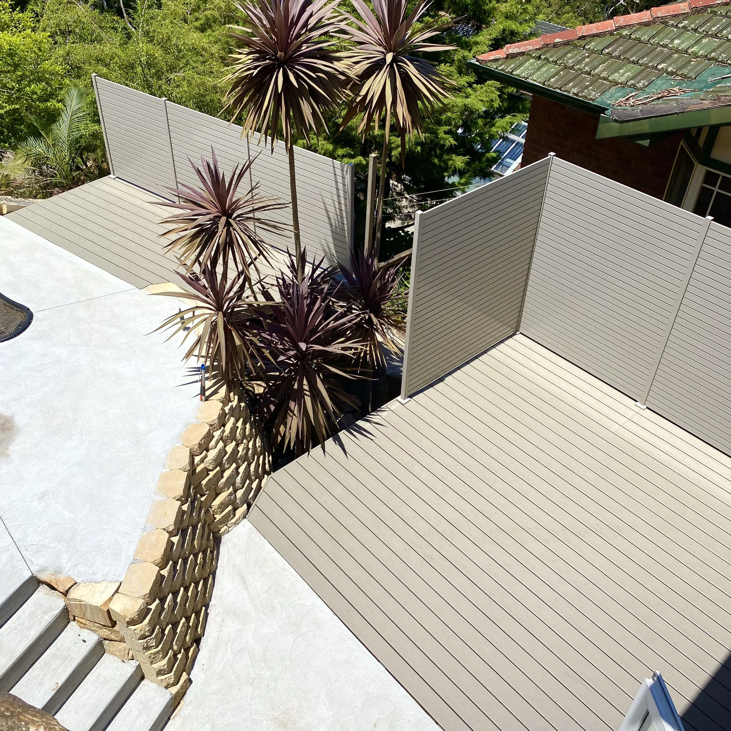 View of a backyard patio area with composite deck, a white privacy fence, a stone wall with purple-leafed plants, and part of a house with a green roof. Bought to you by Platinum Decks.