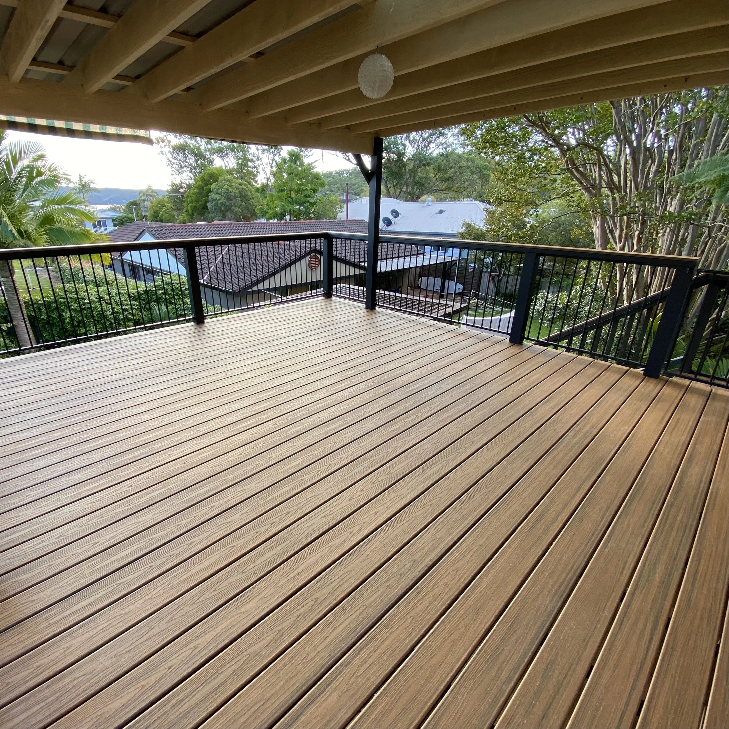 View from a balcony with a brown composite decking and black railing, overlooking rooftops and trees with a backdrop of distant hills and a clear sky.