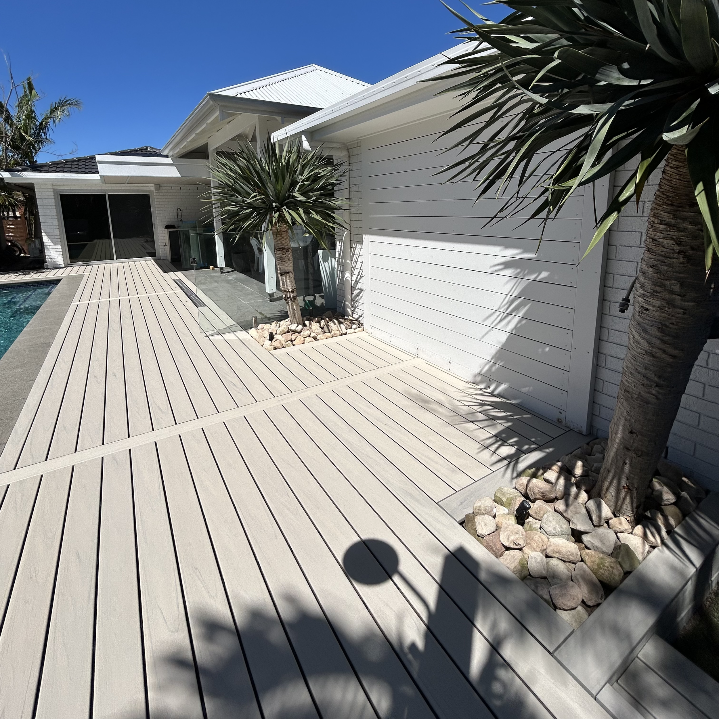 Trex Salt Ash composite deck by Platinum Decks & Patios, creating a light coastal backyard deck in front of a white exterior house, a metal roof, and large sliding glass doors, with desert plants and trees in the foreground, against a clear blue sky.