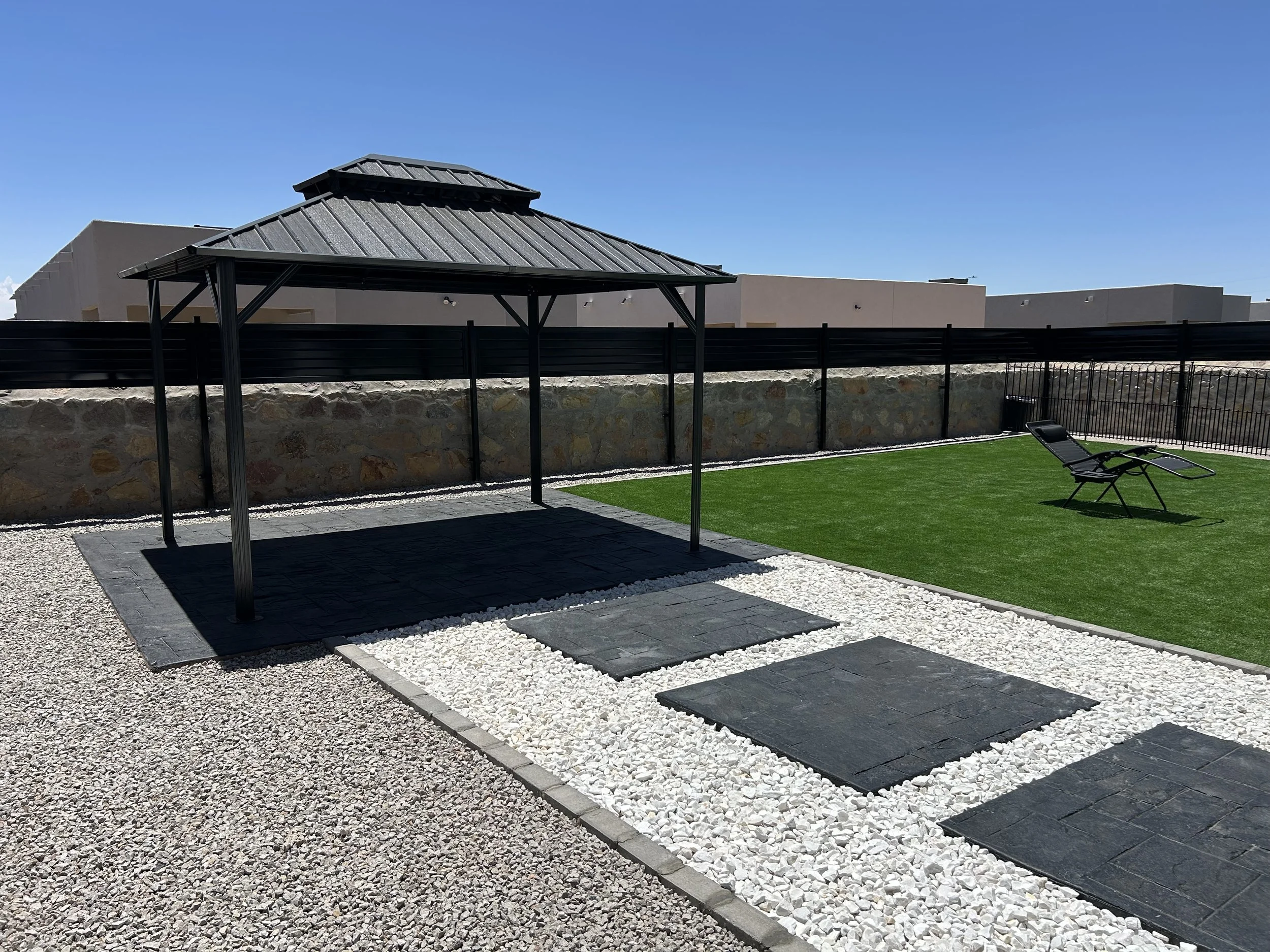 A backyard with a stone wall topped by a black fence, a grass area with a lounge chair, a covered patio with stone pavers, and a modern house with a metal roof under a clear blue sky.