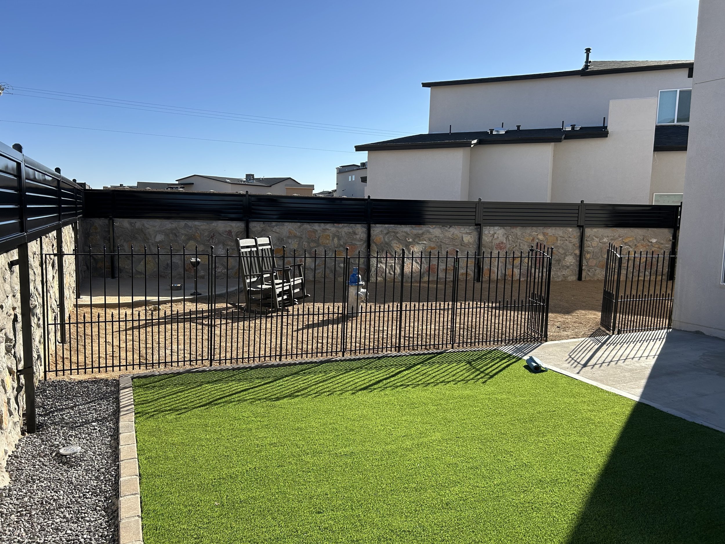 A backyard with a green lawn, a stone wall, a black metal fence, a seating area with chairs, and neighboring white buildings under a clear blue sky.