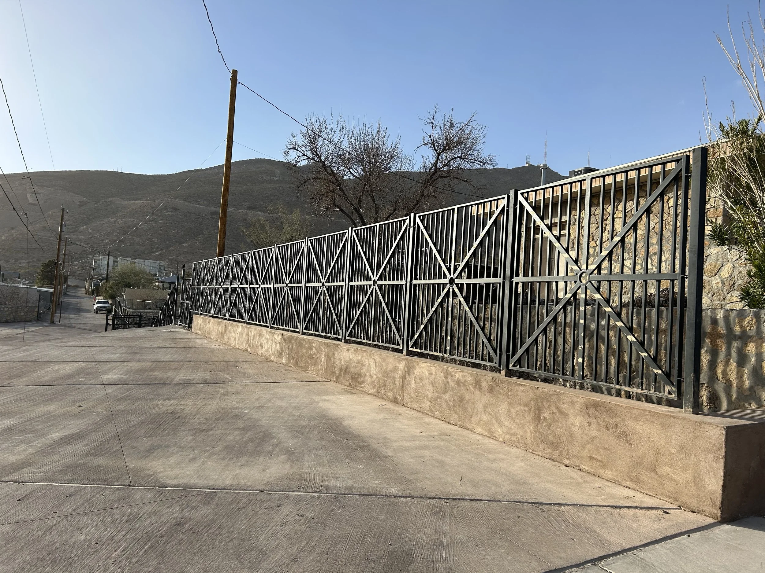 A paved sidewalk with a metal fence and a stone wall, dry leafless trees, power lines, a hill in the background, and a clear blue sky.