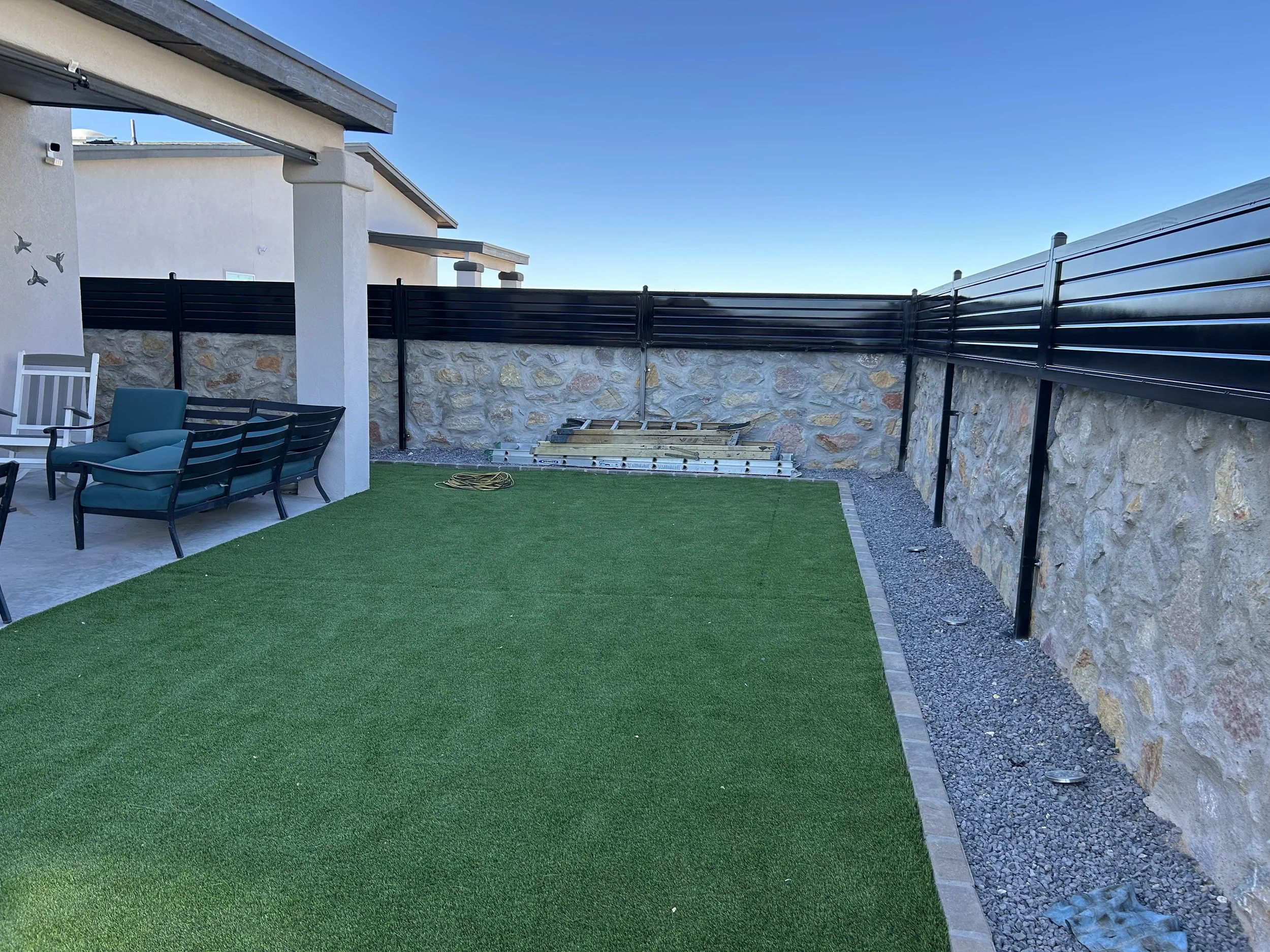 Backyard with a dark fence, stone wall, patio furniture, artificial grass, and a clear blue sky.