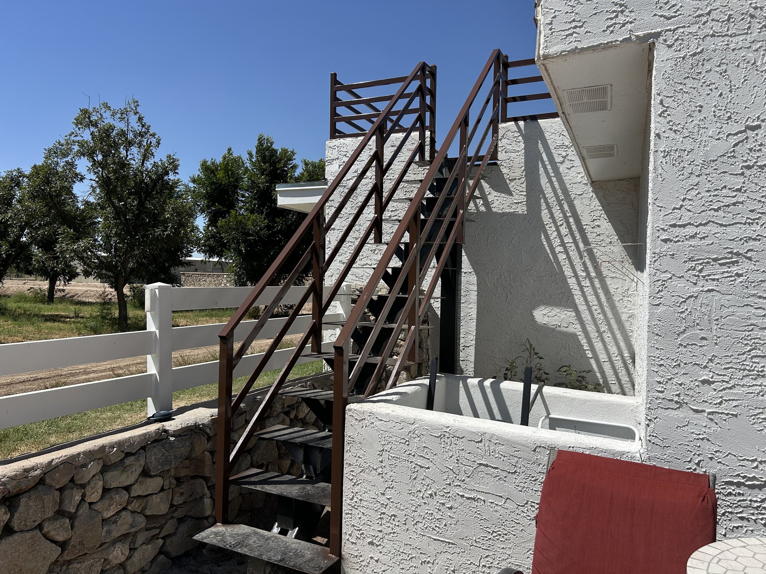 An exterior view of a white stucco building with brown metal stairs leading up to a small balcony with a brown railing. There are trees and a clear blue sky in the background.