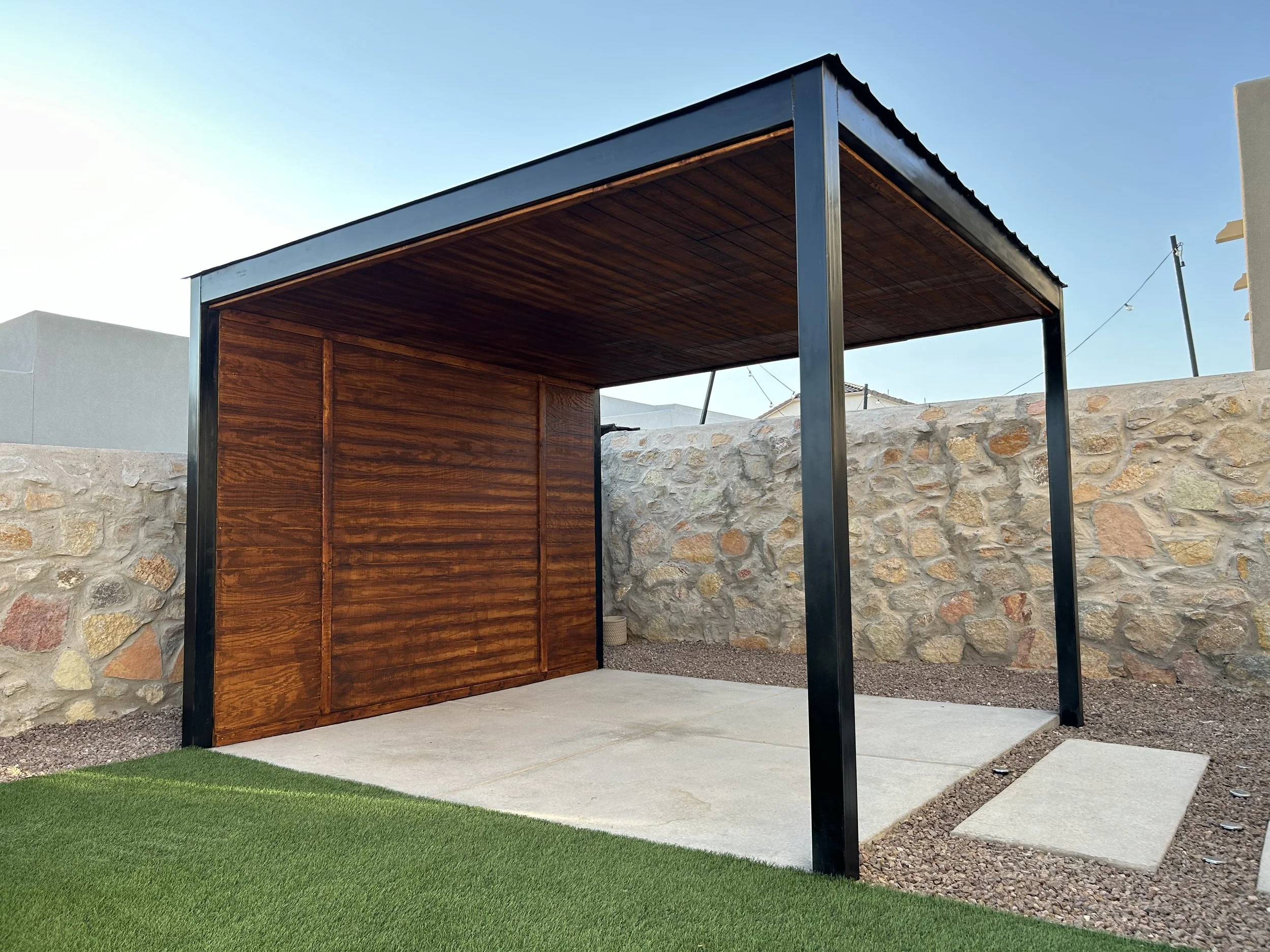 Modern outdoor structure with a wooden wall, black metal frame, concrete floor, and artificial grass nearby, against a stone wall and clear sky.