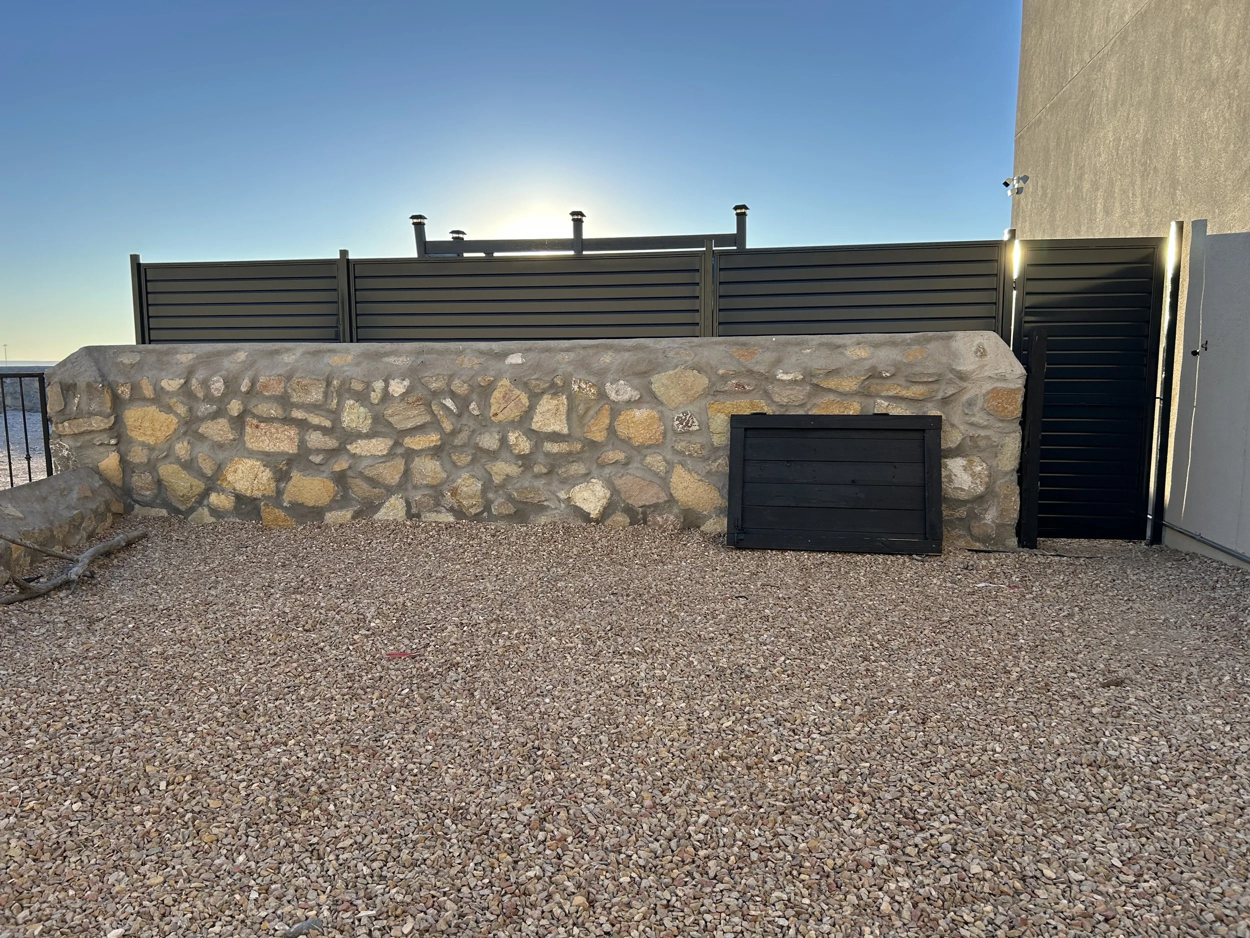 View of a desert landscape with a stone wall, metal gate, and gravel ground. There is a tall fence and security lights on a building to the right. The sky is clear and blue with the sun low on the horizon.
