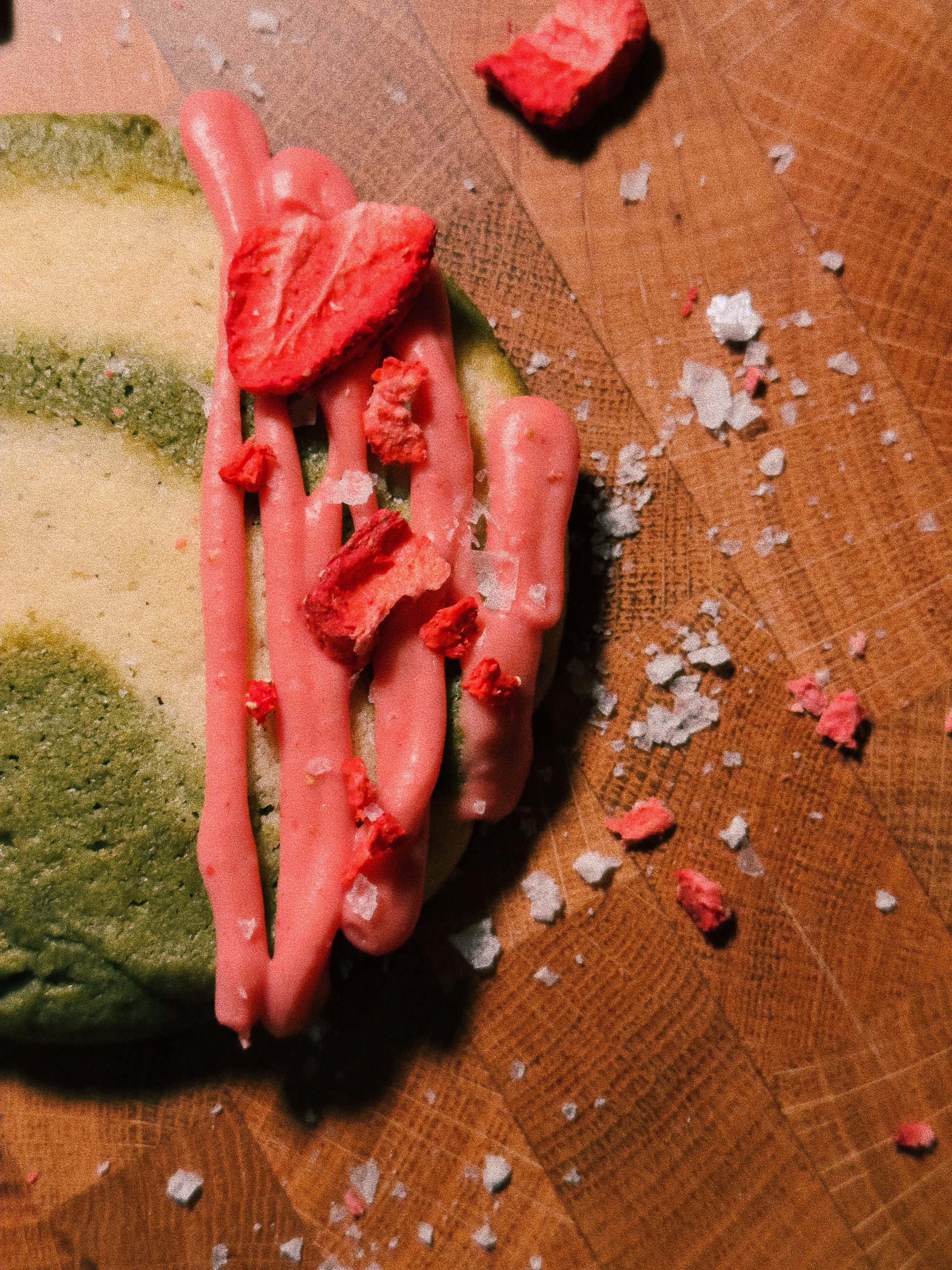 A partially eaten slice of watermelon with pink icing on a wooden surface, surrounded by scattered white and pink crumbs.