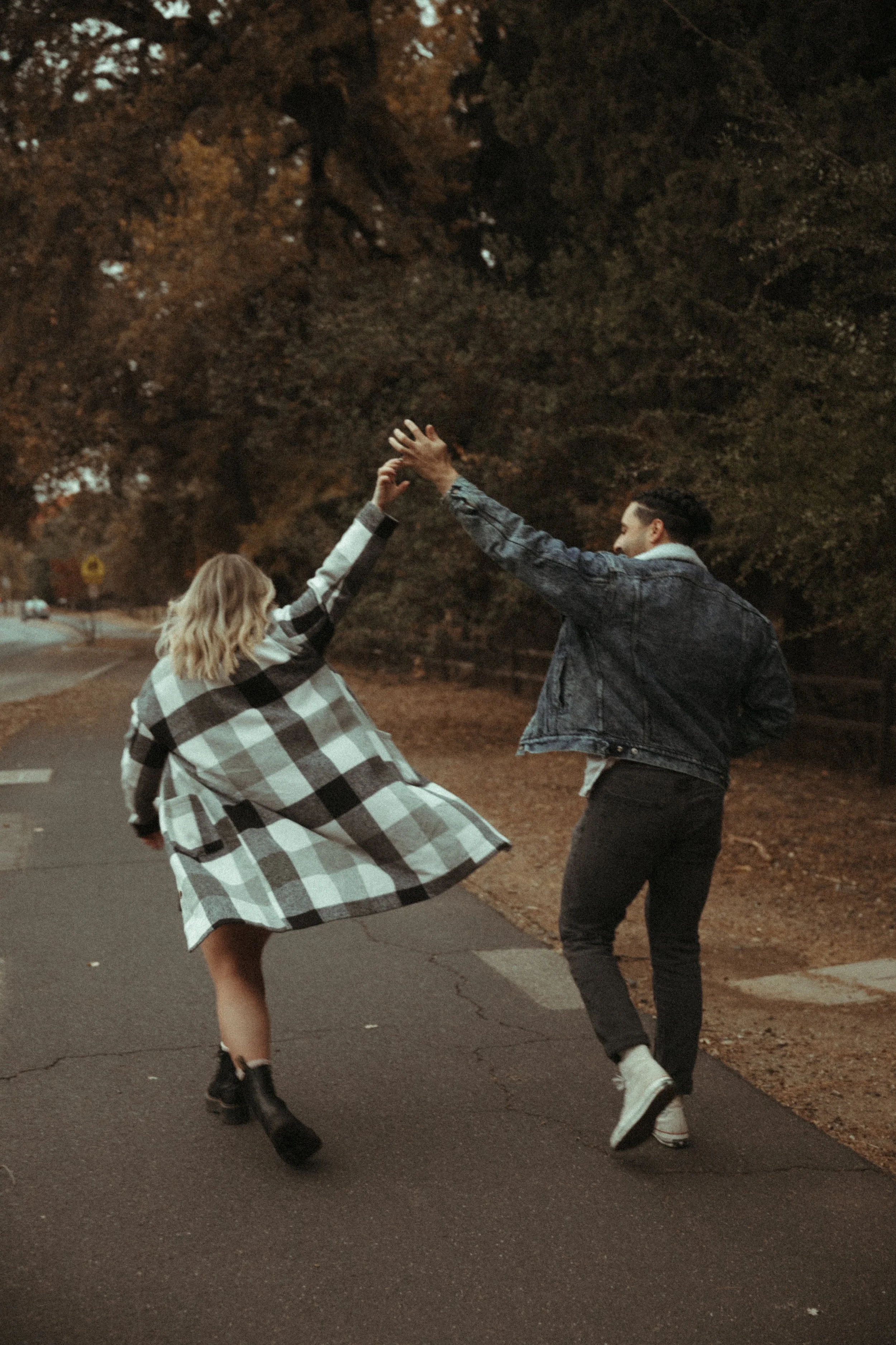 A young woman and a young man dancing outdoors at dusk. The woman is wearing a black and white checkered coat and boots, and the man is wearing a denim jacket and sneakers. They are holding hands and dancing near a tree line.