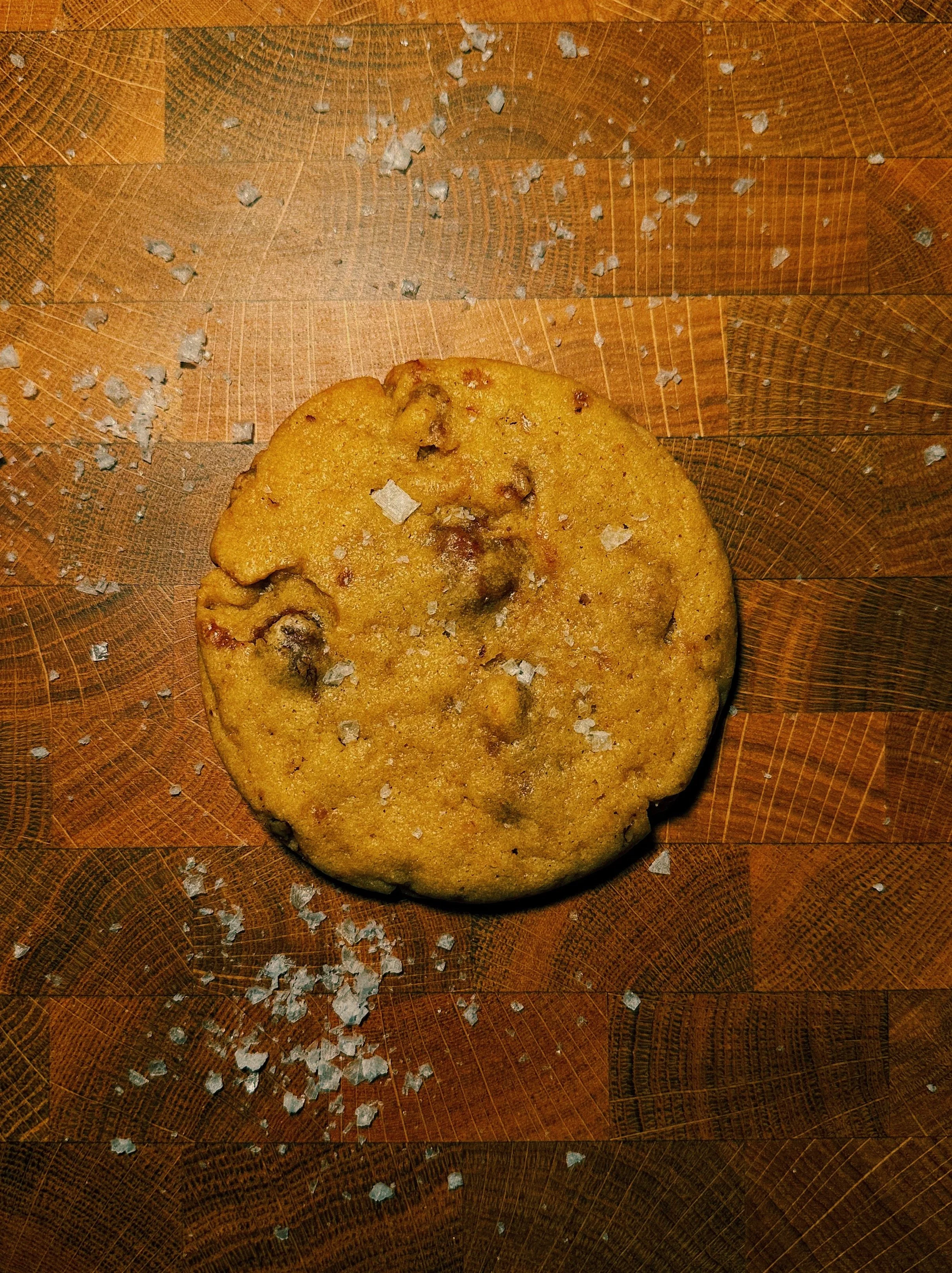Close-up of a chocolate chip cookie sprinkled with salt on a wooden cutting board with salt scattered around.