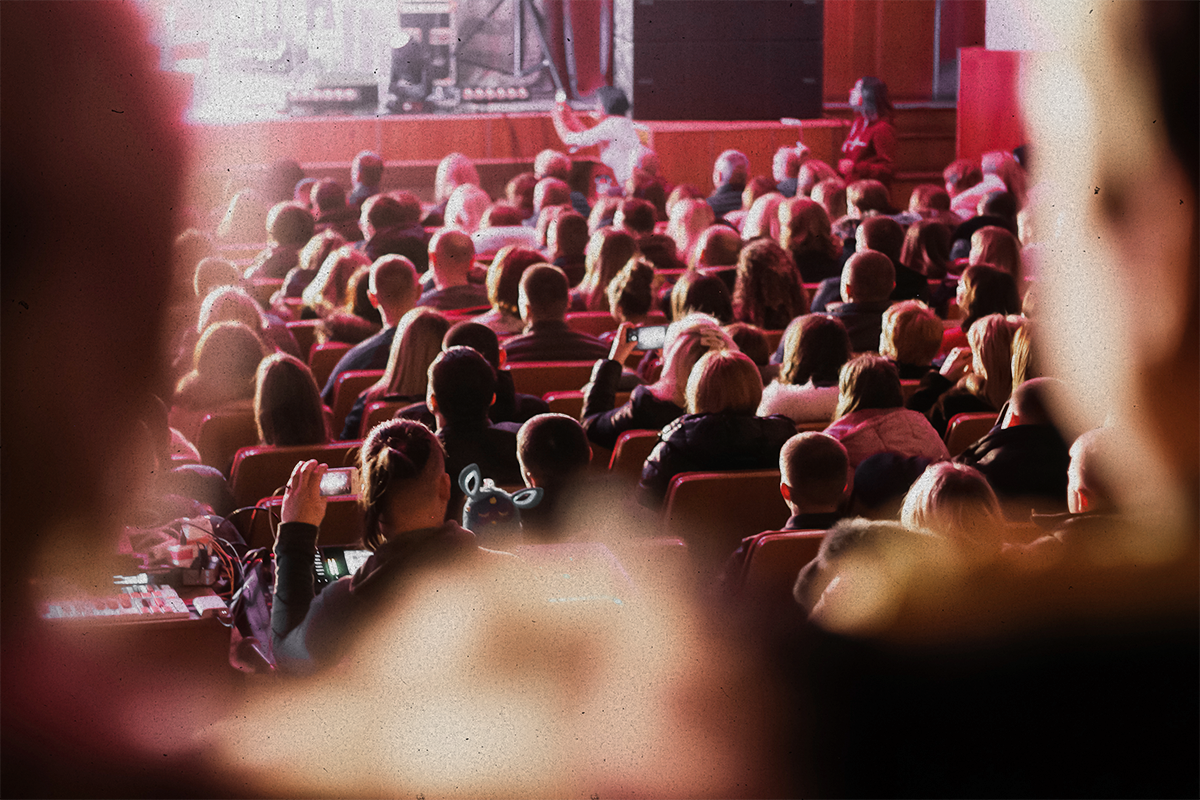 Film audience seated in a theater with warm red lighting, viewed from behind — representing the Bay Area film community gathering for a screening or event.