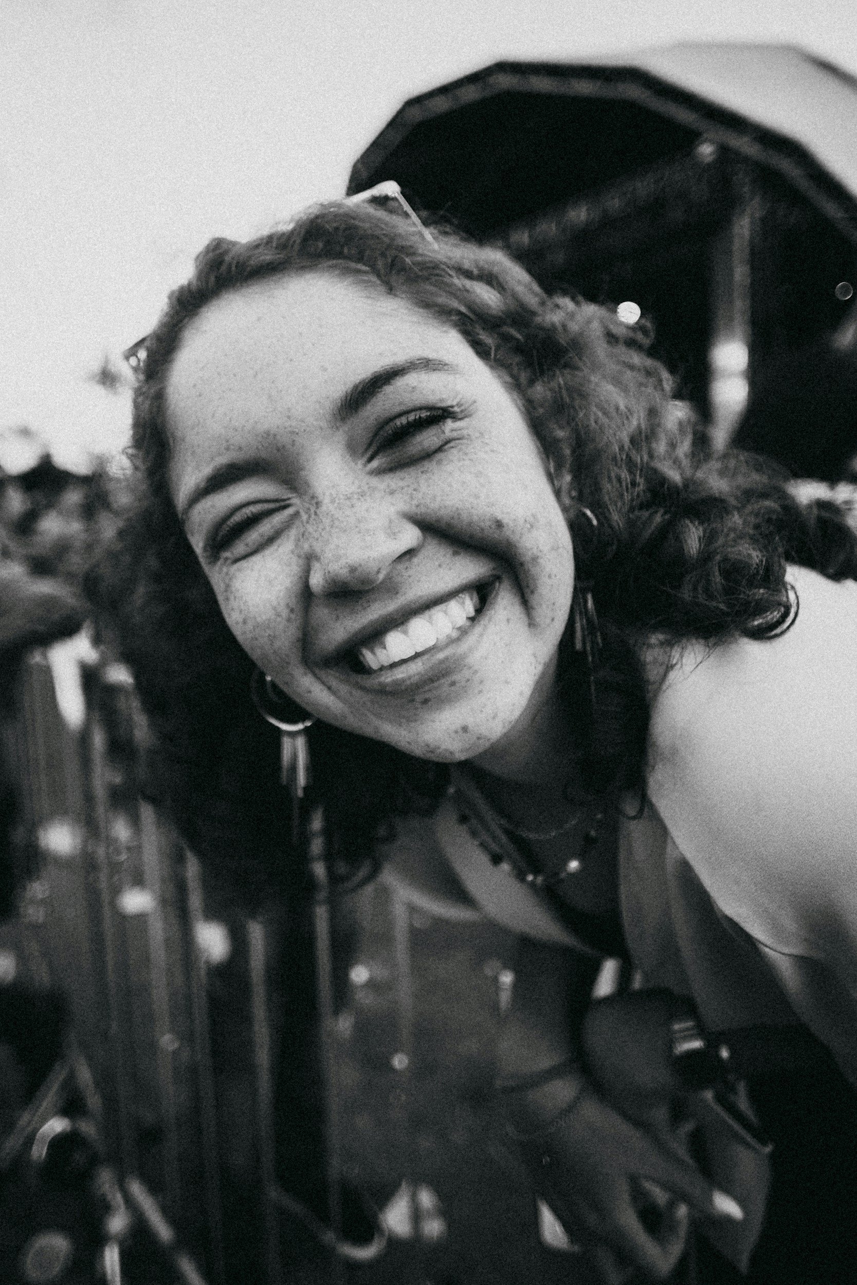 Close-up of a happy woman with curly hair, taking a selfie, with a blurred background of structures, in black and white.