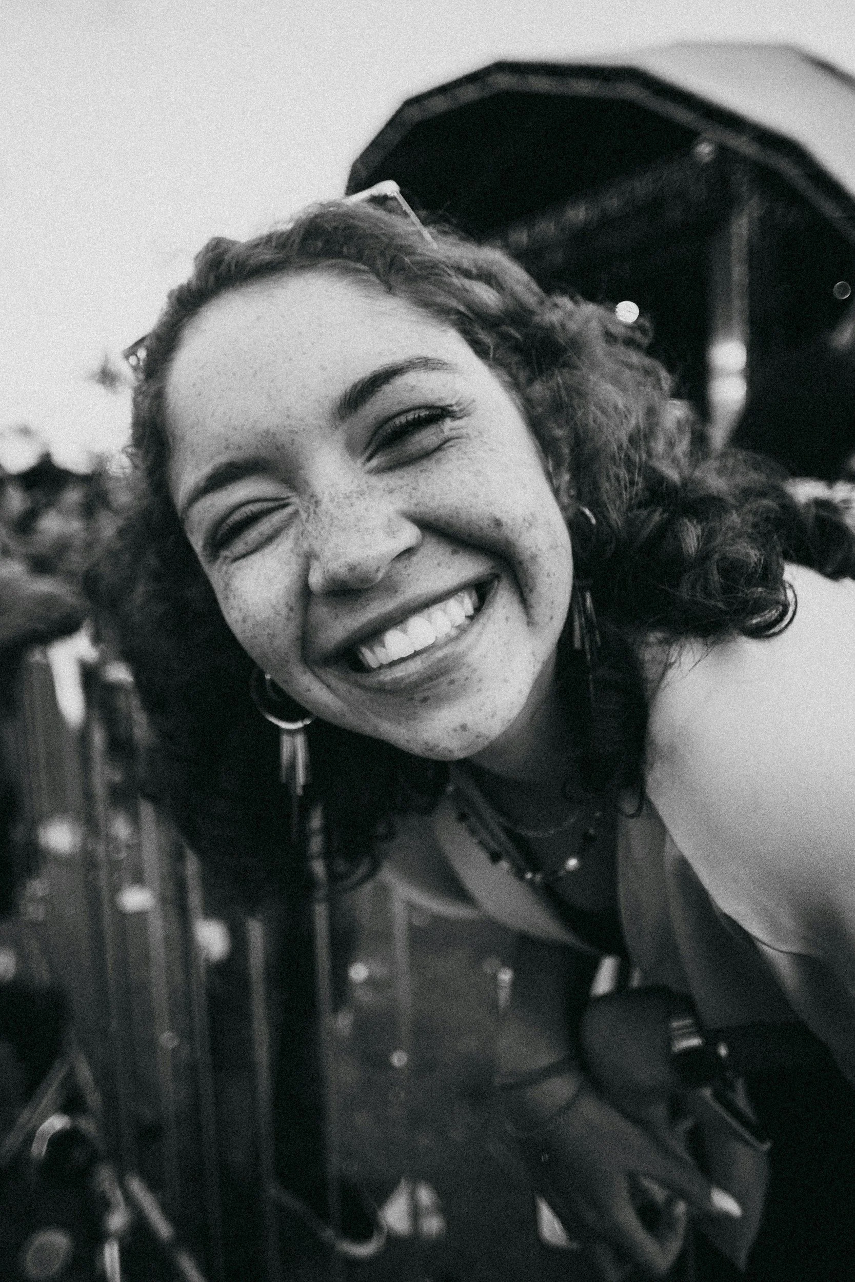 Close-up of a smiling woman with curly hair and freckles, wearing sunglasses on her head, leaning on a balcony railing with blurred city lights in the background.