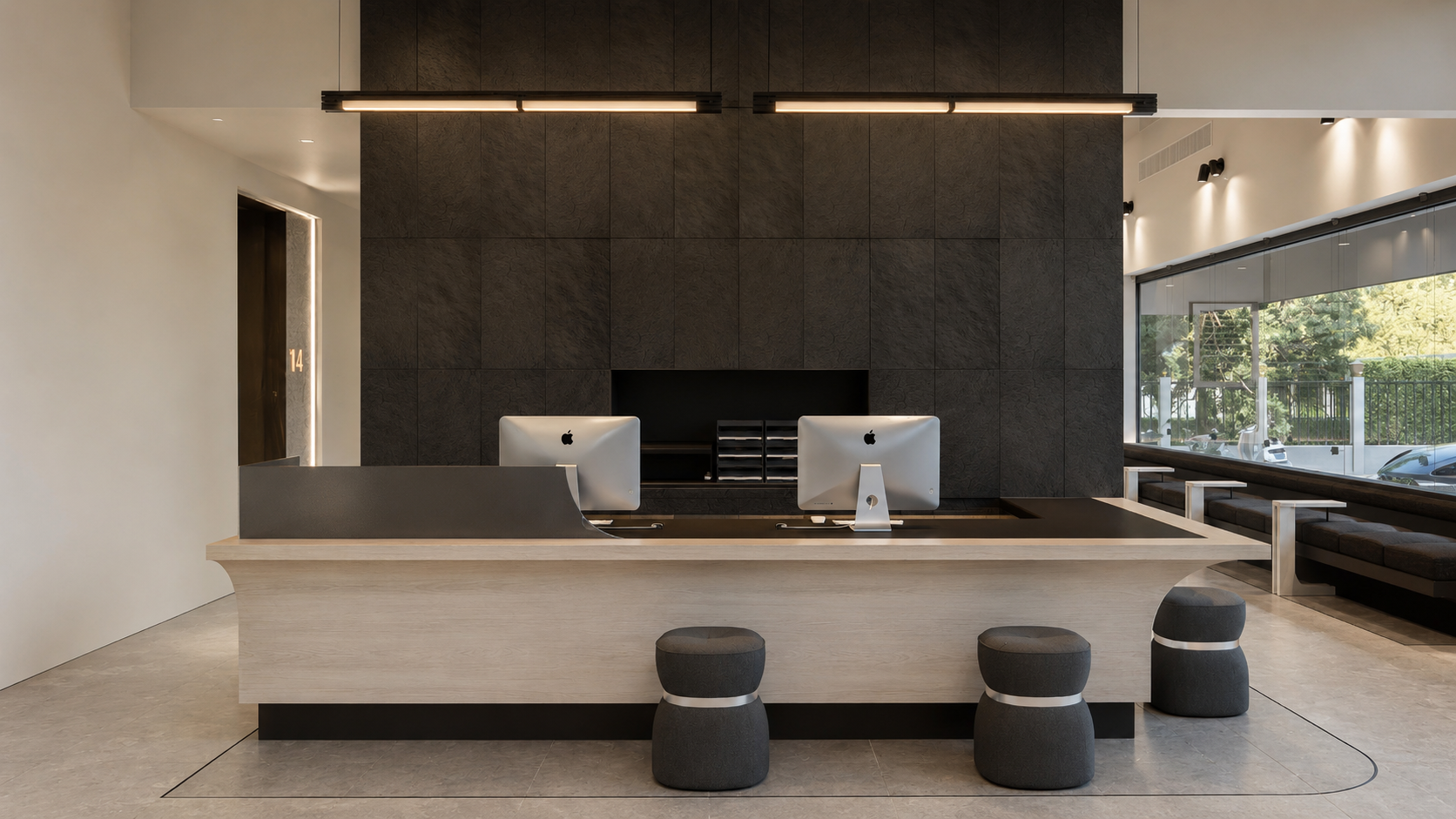 Modern reception area with a light wooden counter, two Apple iMac computers, black side panels, dark textured wall behind, large window on the right, and seating stools in front.