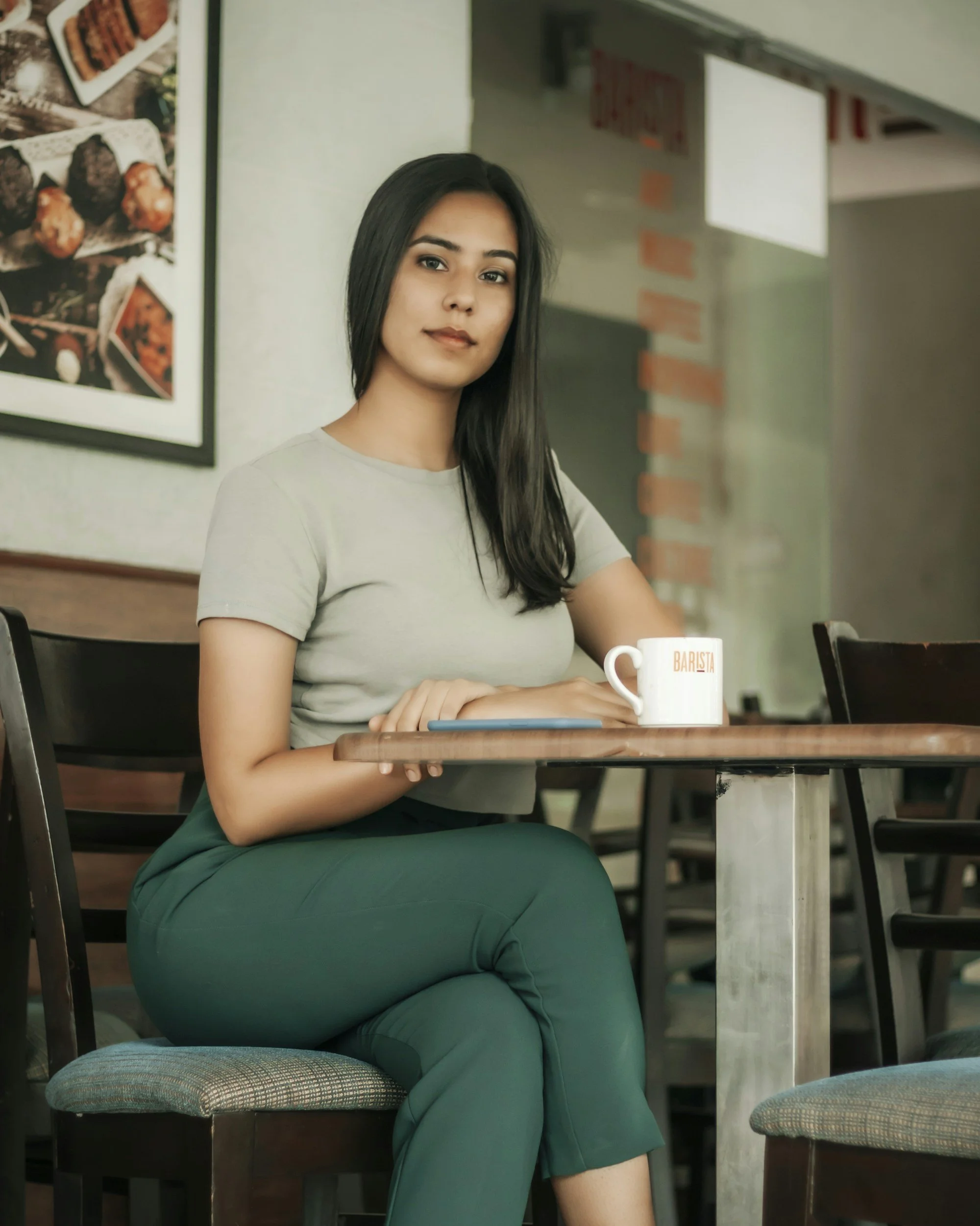 A young woman with long dark hair sitting at a dining table inside a cafe or restaurant, with a white coffee mug in front of her. She is wearing a light grey t-shirt and dark green pants, looking at the camera with a slight smile. There are chairs around her and a wall with pictures of food in the background.
