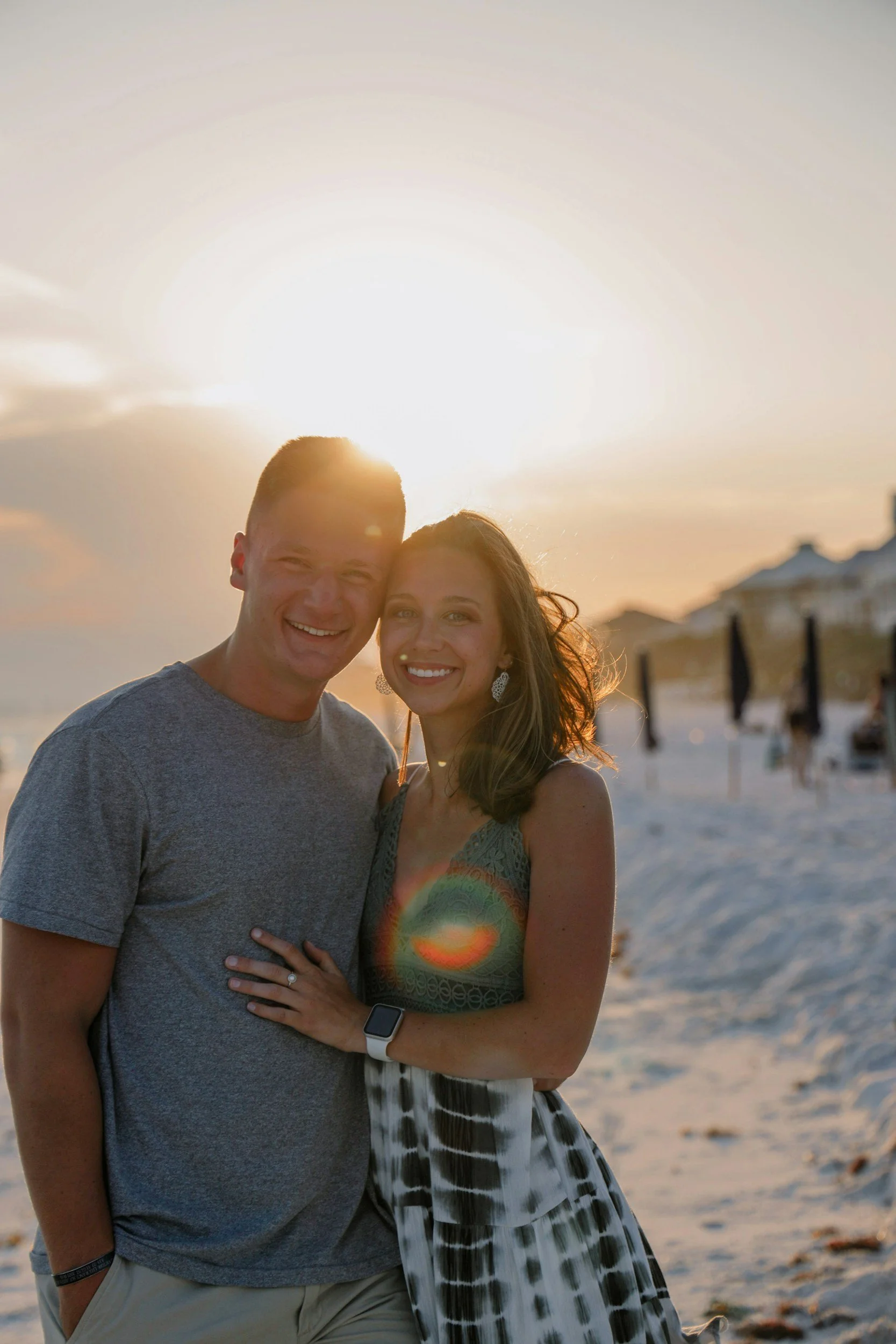 A smiling couple standing close together on a beach at sunset, with the sun behind them and a few clouds in the sky.