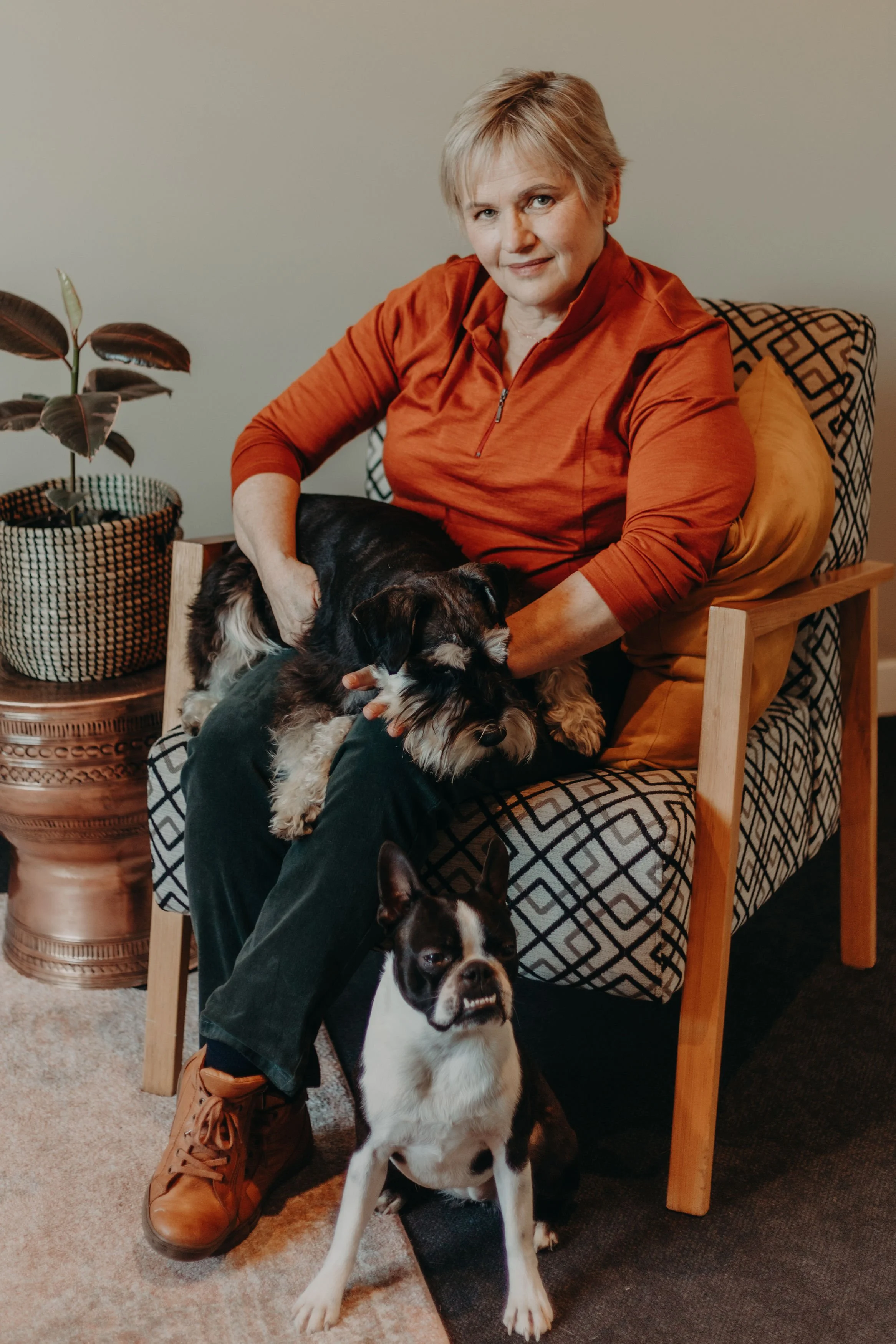 A woman with short blonde hair and a burnt orange zip-up jacket sitting on a patterned armchair, holding a small black and white puppy. A large potted plant is beside her, and a black and white dog is sitting on the floor in front.