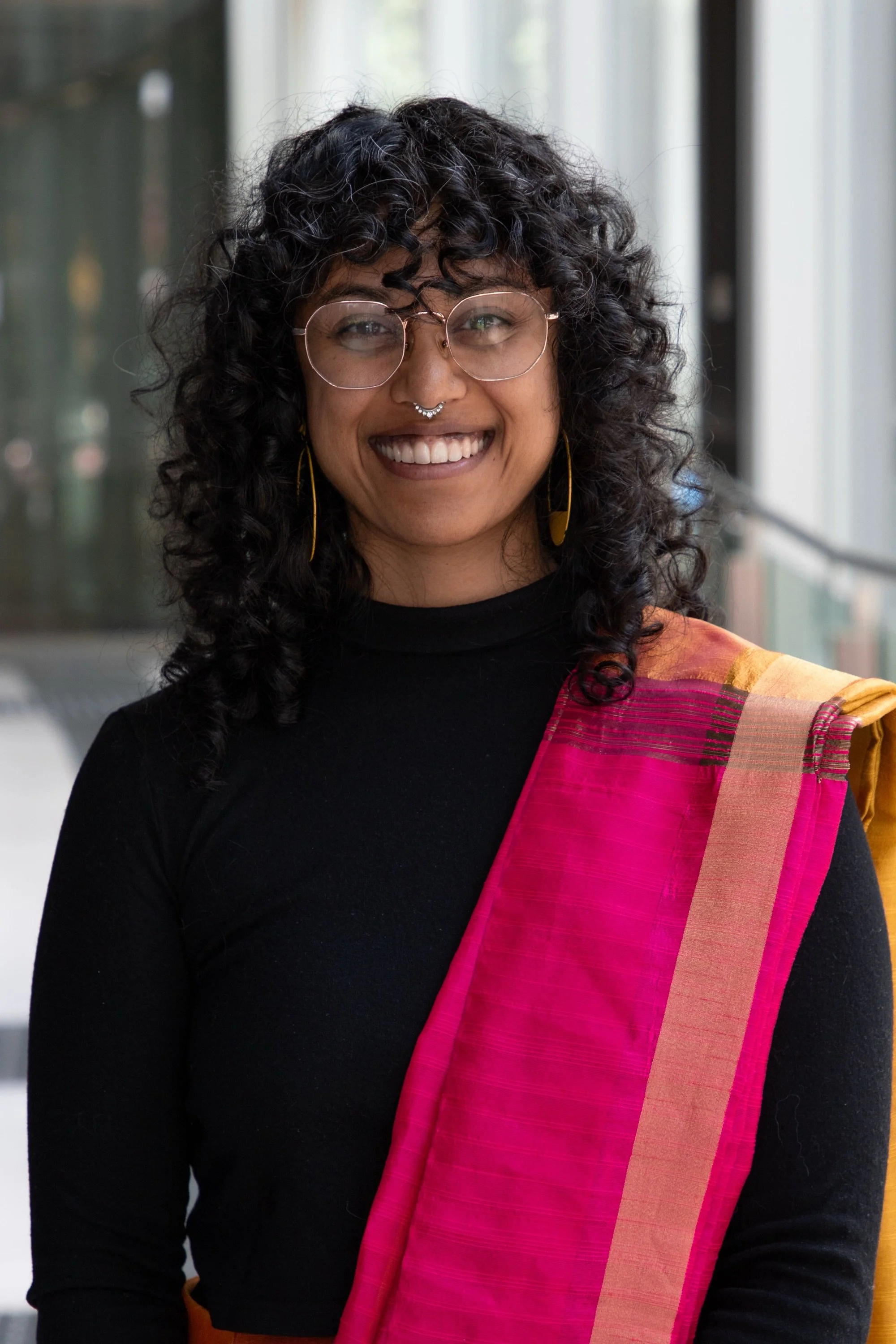 A woman with curly black hair, glasses, and a septum piercing smiling at the camera, wearing a traditional colorful sari with a black top indoors.