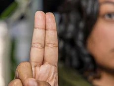 Close-up of person's hand showing three fingers raised, with part of face visible in background