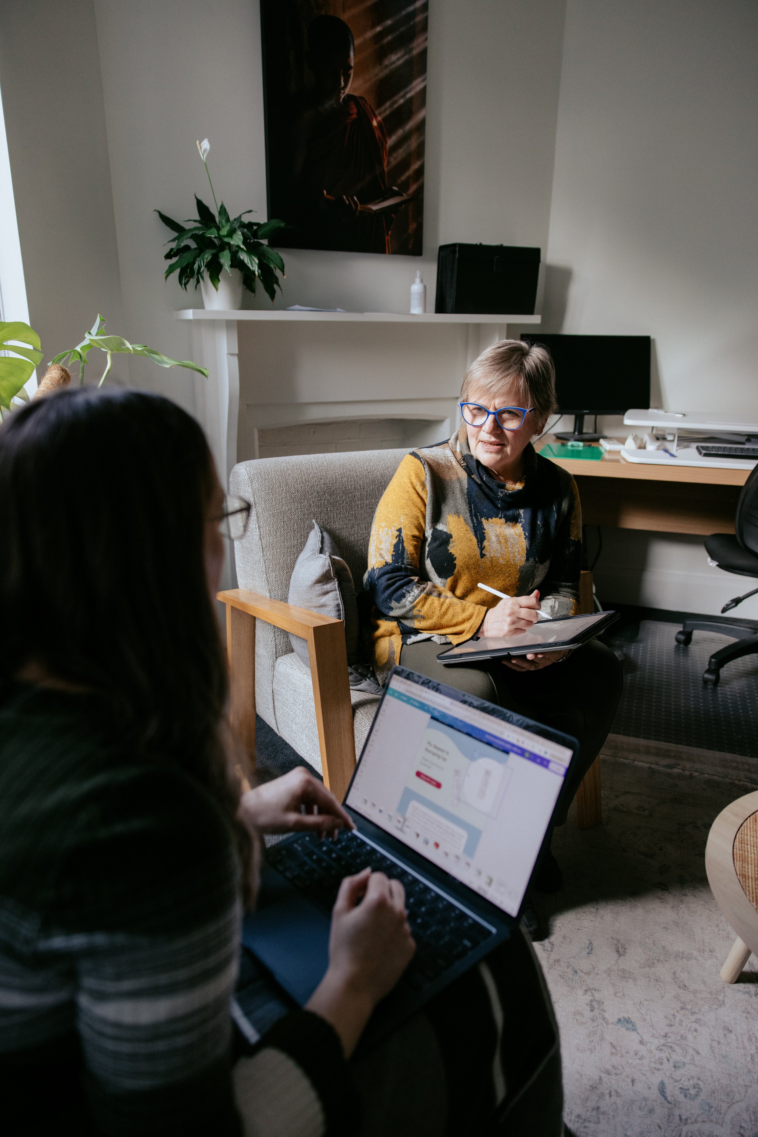 Women in a meeting discussing work, with a woman in glasses taking notes and others using laptops in an office setting.