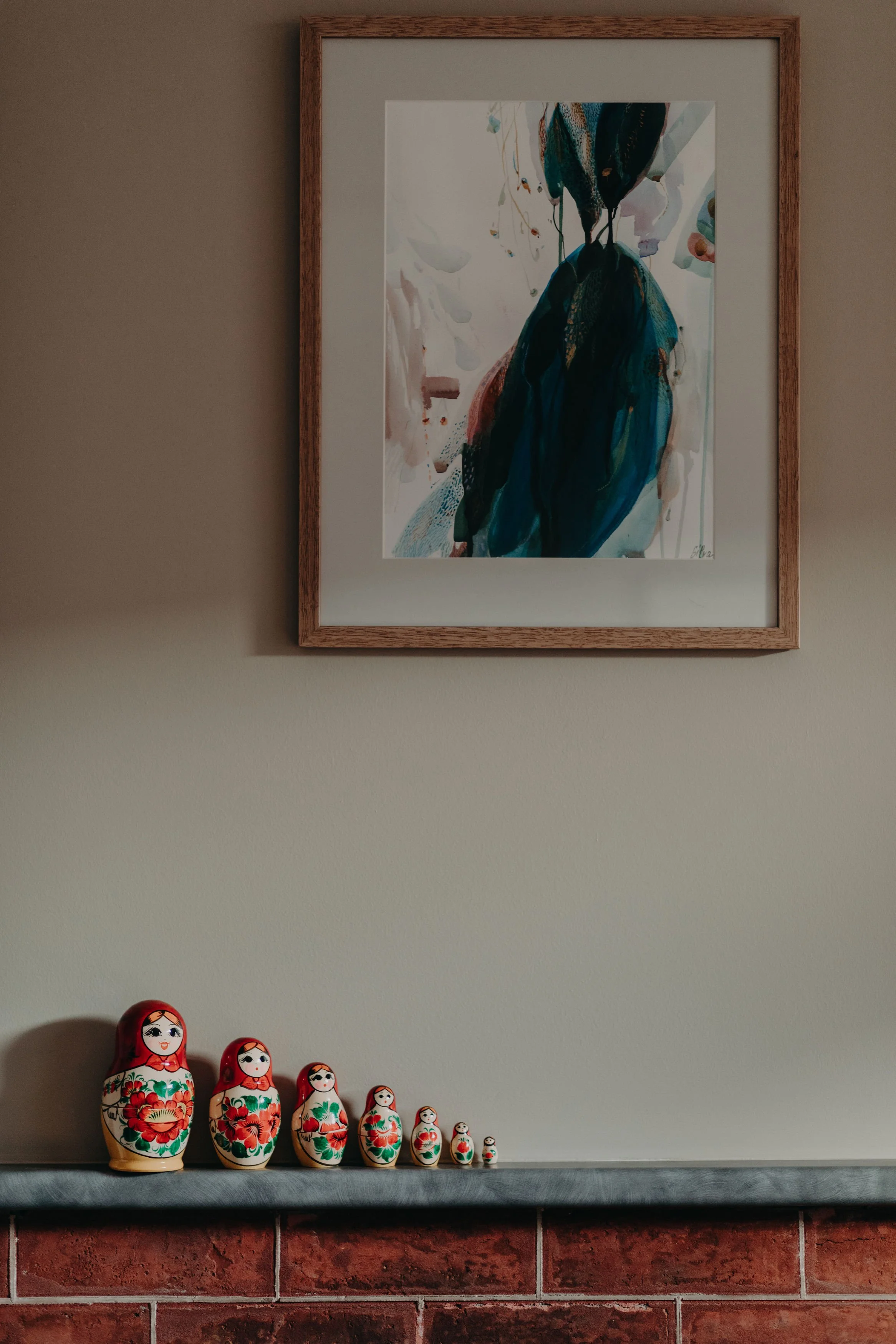 A row of Russian nesting dolls, called Matryoshka dolls, of varying sizes with red and green floral designs, displayed on a ledge against a brick wall. Above them, a framed watercolor painting of a bird in blue and black tones hangs on a beige wall.
