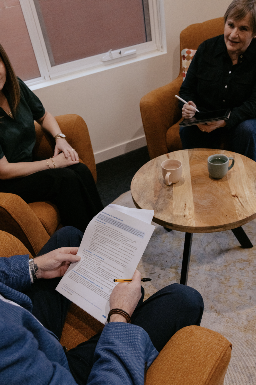 Three people sitting in a cozy room having a discussion. One person is holding papers and a pen, another is holding a digital tablet and a stylus, and the third is sitting with their hands folded. There are cups on a wooden coffee table and a window in the background.