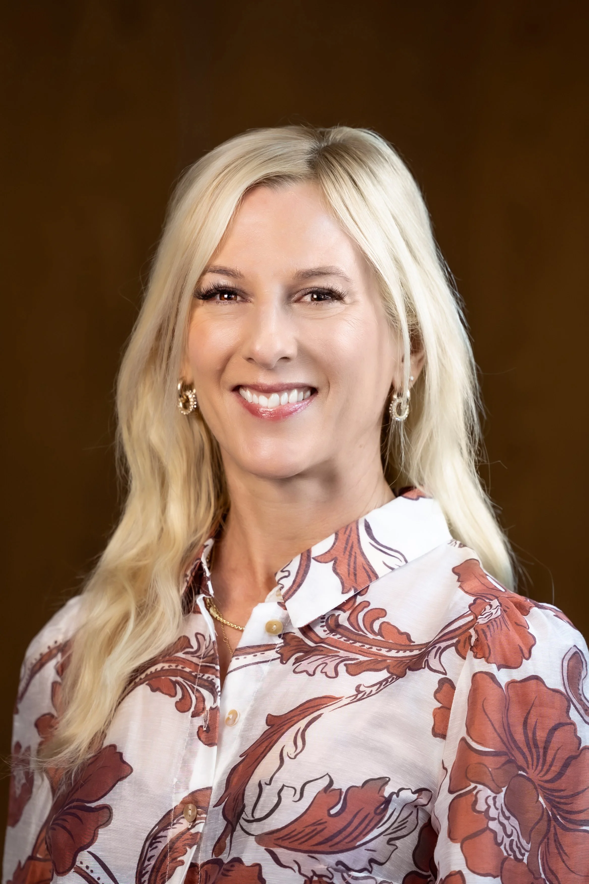 A smiling blonde woman with long wavy hair wearing a patterned button-up shirt with floral designs, earrings, and a necklace.