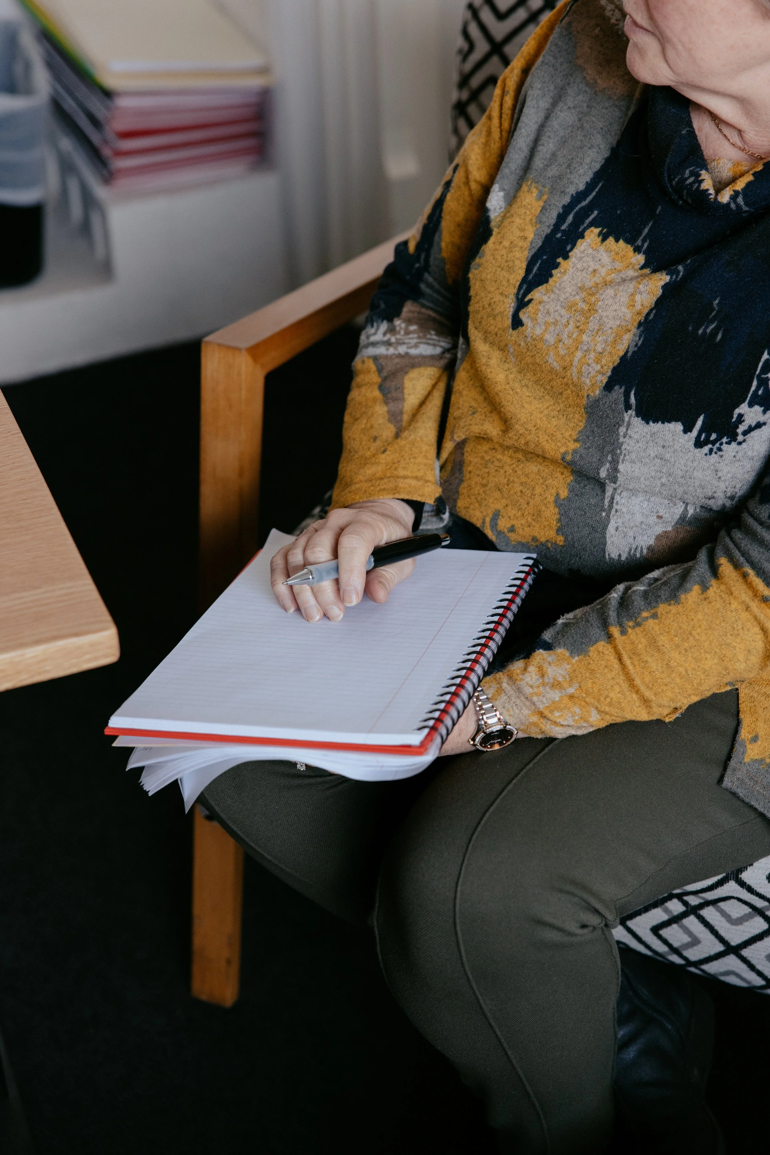 A person sitting on a chair, holding a spiral notebook and a pen, with a partial view of their face, wearing a mustard, gray, and black patterned sweater. There are stacks of papers and folders on a shelf in the background.