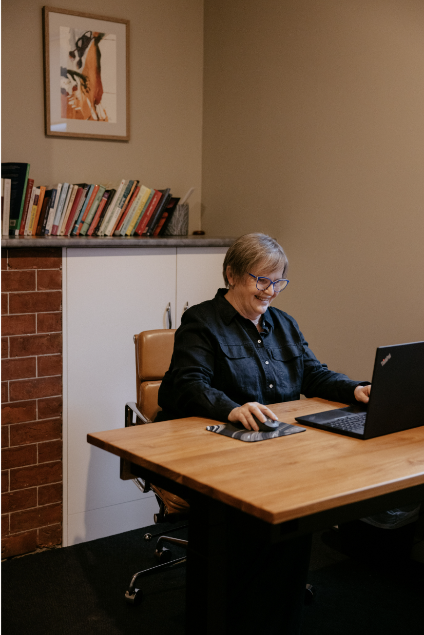 A woman with short hair and glasses sitting at a wooden desk with a laptop and computer mouse, smiling in a home office or study room with a bookshelf and framed artwork on the wall.