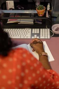 A person with curly hair wearing an orange polka dot shirt writing on a piece of paper at a desk with a keyboard, potted plant, and pink bowl nearby.