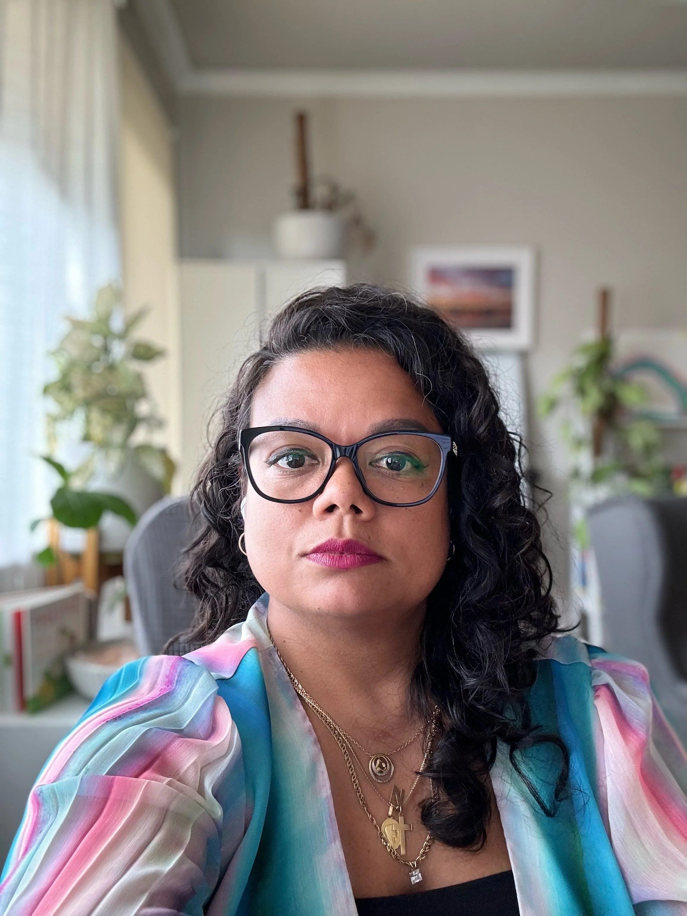 A woman with dark curly hair and glasses, wearing a multicolored, pastel, tie-dye shirt, sitting in an indoor space with plants and artwork in the background.