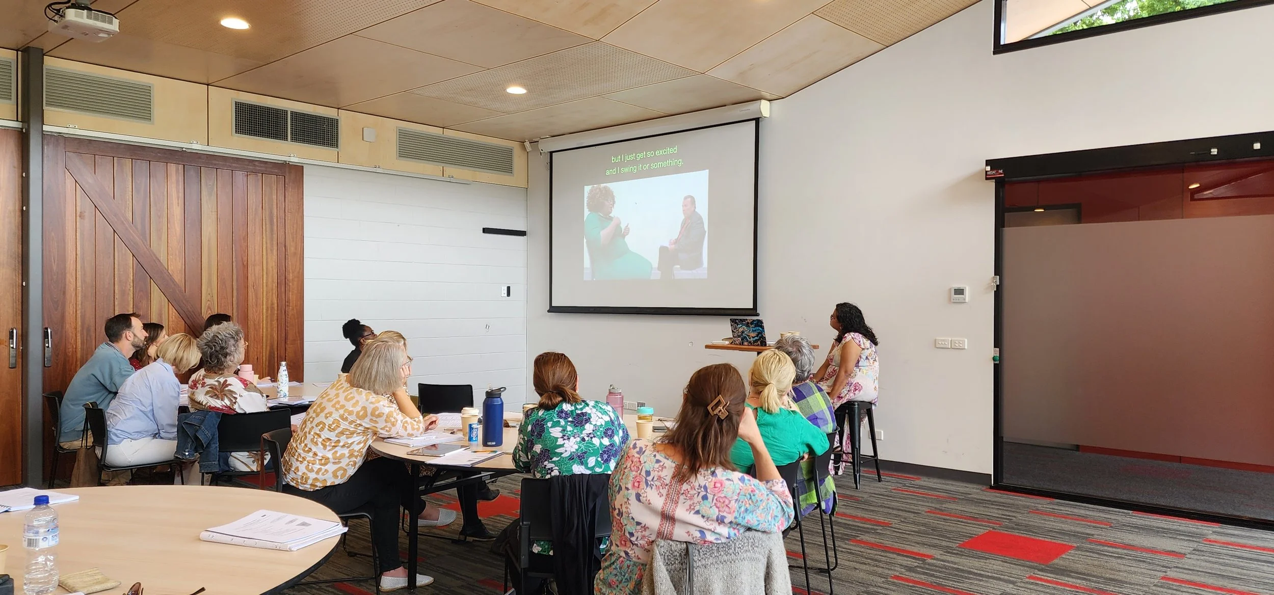 A group of people attending a presentation or workshop in a modern conference room. The attendees are seated at tables, watching a speaker standing near a large screen displaying a slide with text and images.