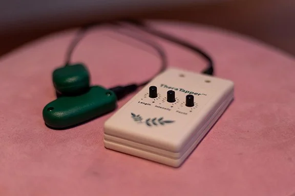 A small white electronic device with three black knobs and some decorative black markings, connected to green earbuds on a pink surface.