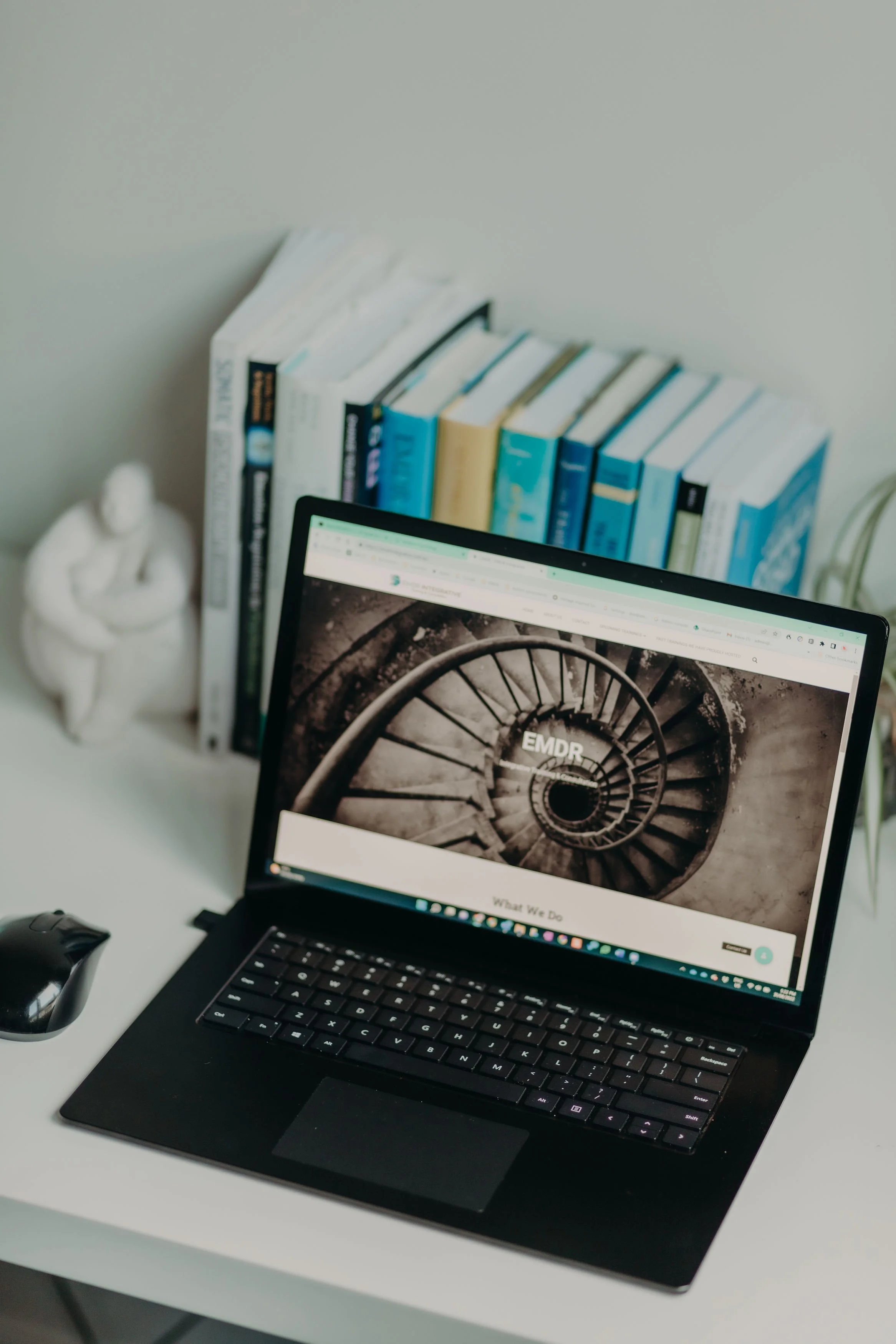 A laptop on a white desk with a webpage open showing a spiral staircase on the screen. Behind the laptop, there are several stacked books, a small sculpture, and a wire-frame container. A mouse is placed next to the laptop on the desk.