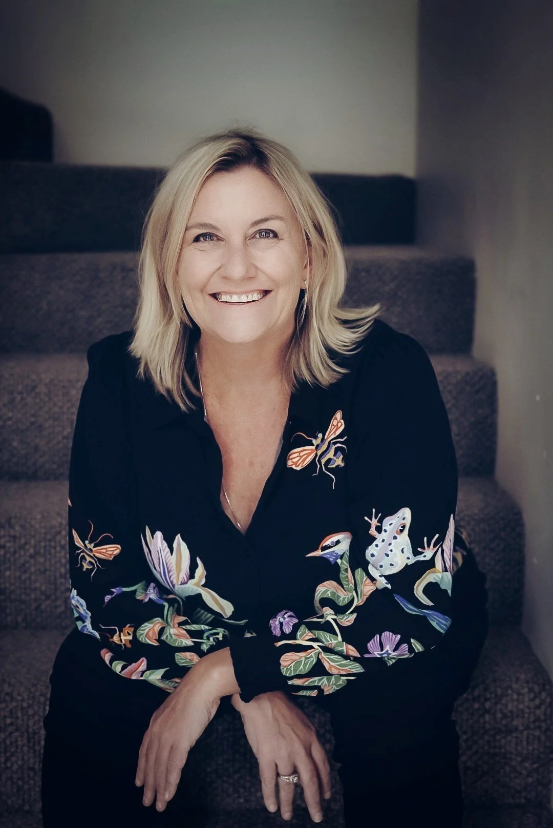 A smiling woman with blonde hair, wearing a black blouse with colorful embroidery of flowers, frogs, and butterflies, sitting on carpeted stairs.