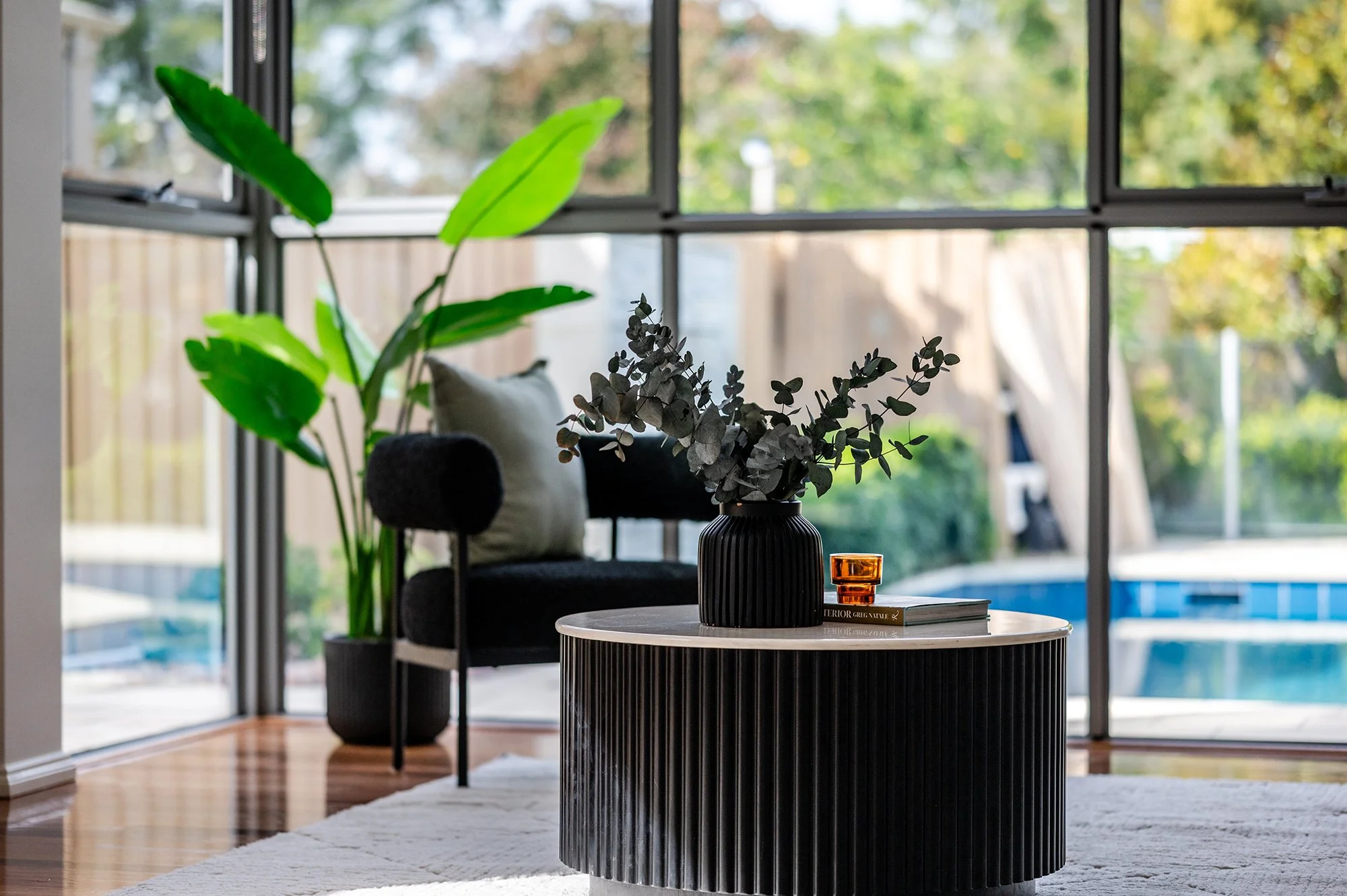 Modern living room with black and white round coffee table, black vase with green and gray leafy plants, orange glass, gray chair, large window with green foliage outside, white rug, wooden floor.