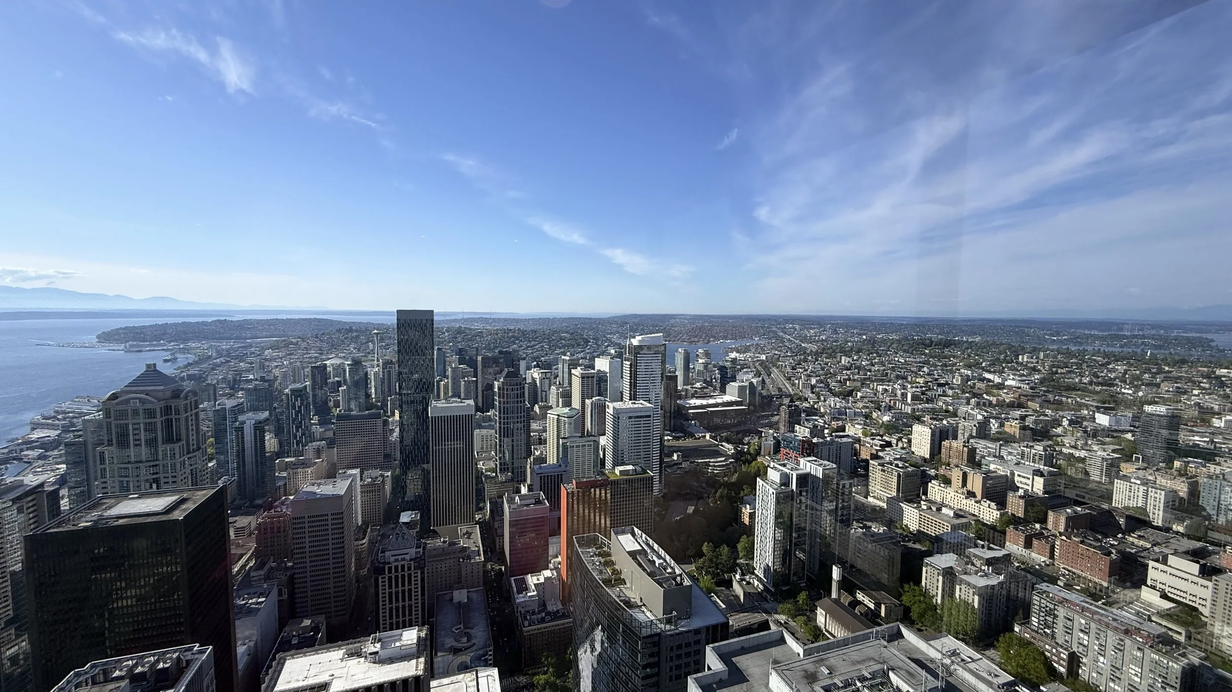 A panoramic view of downtown Seattle from a high vantage point, showing numerous skyscrapers, the Puget Sound, and distant mountains under a partly cloudy sky.