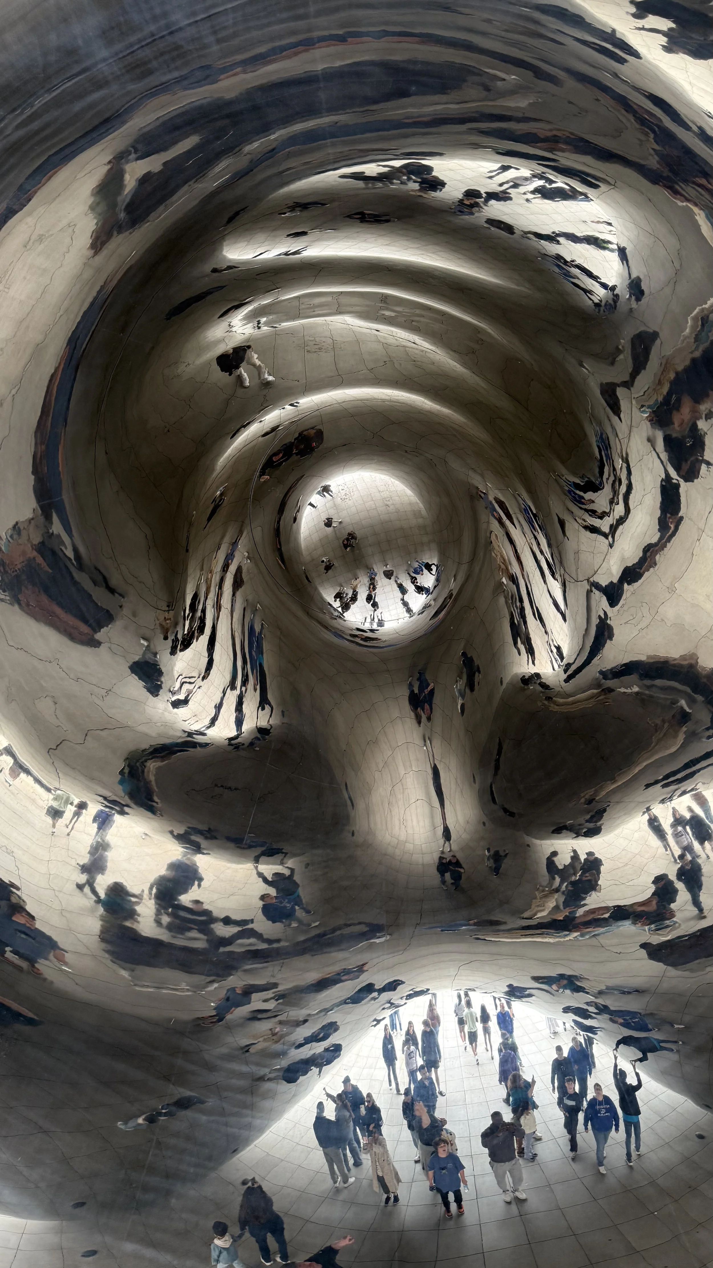 People walking inside a large, reflective, metallic, curved structure, possibly the 'Cloud Gate' sculpture in Chicago, with distorted reflections of visitors.