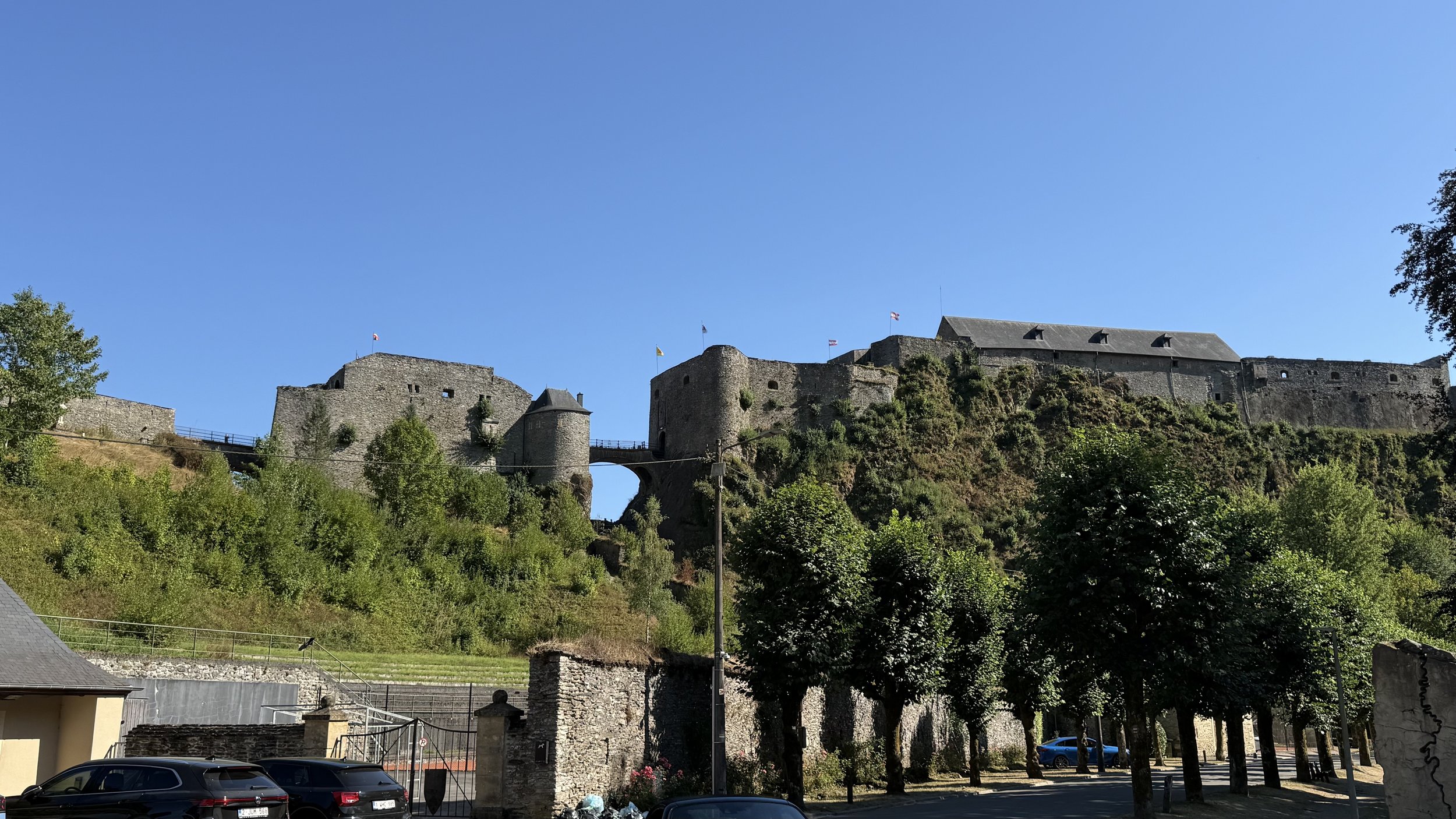A historic stone castle atop a hill with battlements and flags, overlooking a green landscape and parking lot below under a clear blue sky.