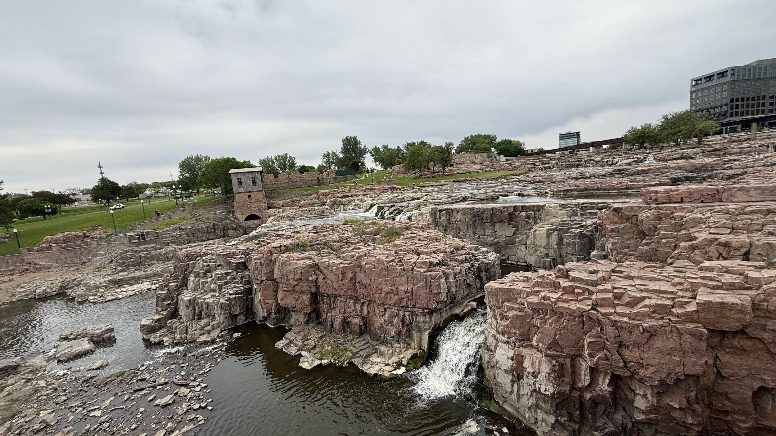 View of a rocky riverbed with small waterfalls, a stone structure, green grass, trees, and modern buildings under a gray cloudy sky.