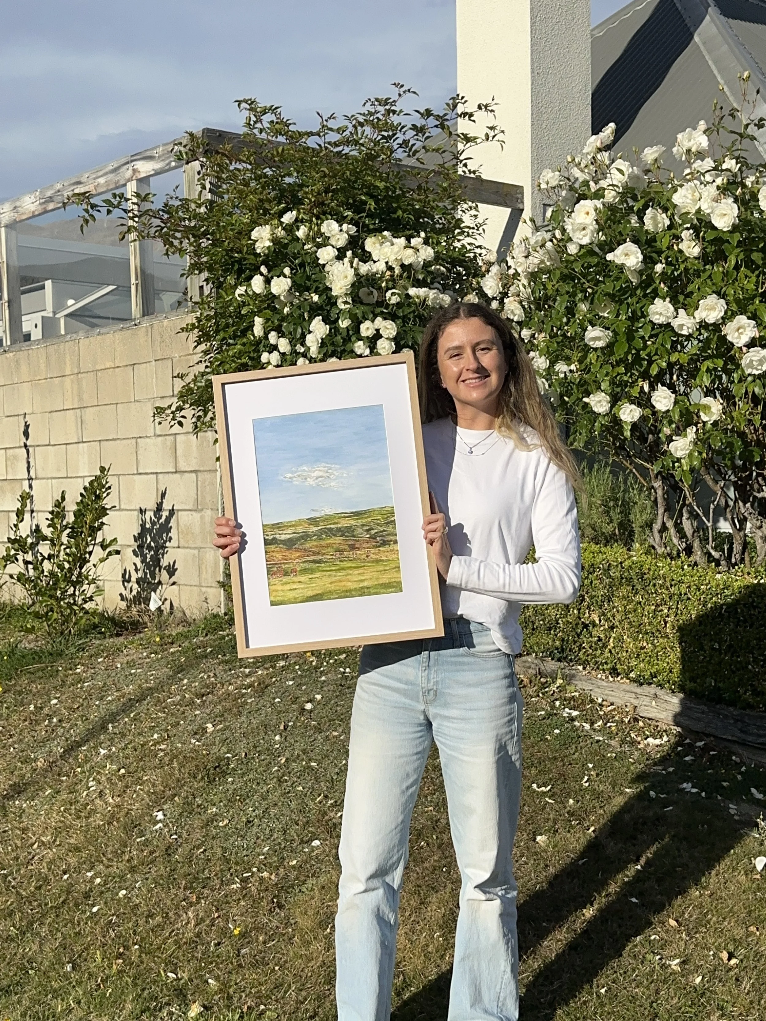 A young woman with long wavy brown hair wearing a white long-sleeve shirt and light blue jeans is smiling and holding a framed landscape painting outdoors. She is standing on grassy ground with small white flowers, in front of a flowering bush with white roses, a brick wall, and modern buildings in the background.