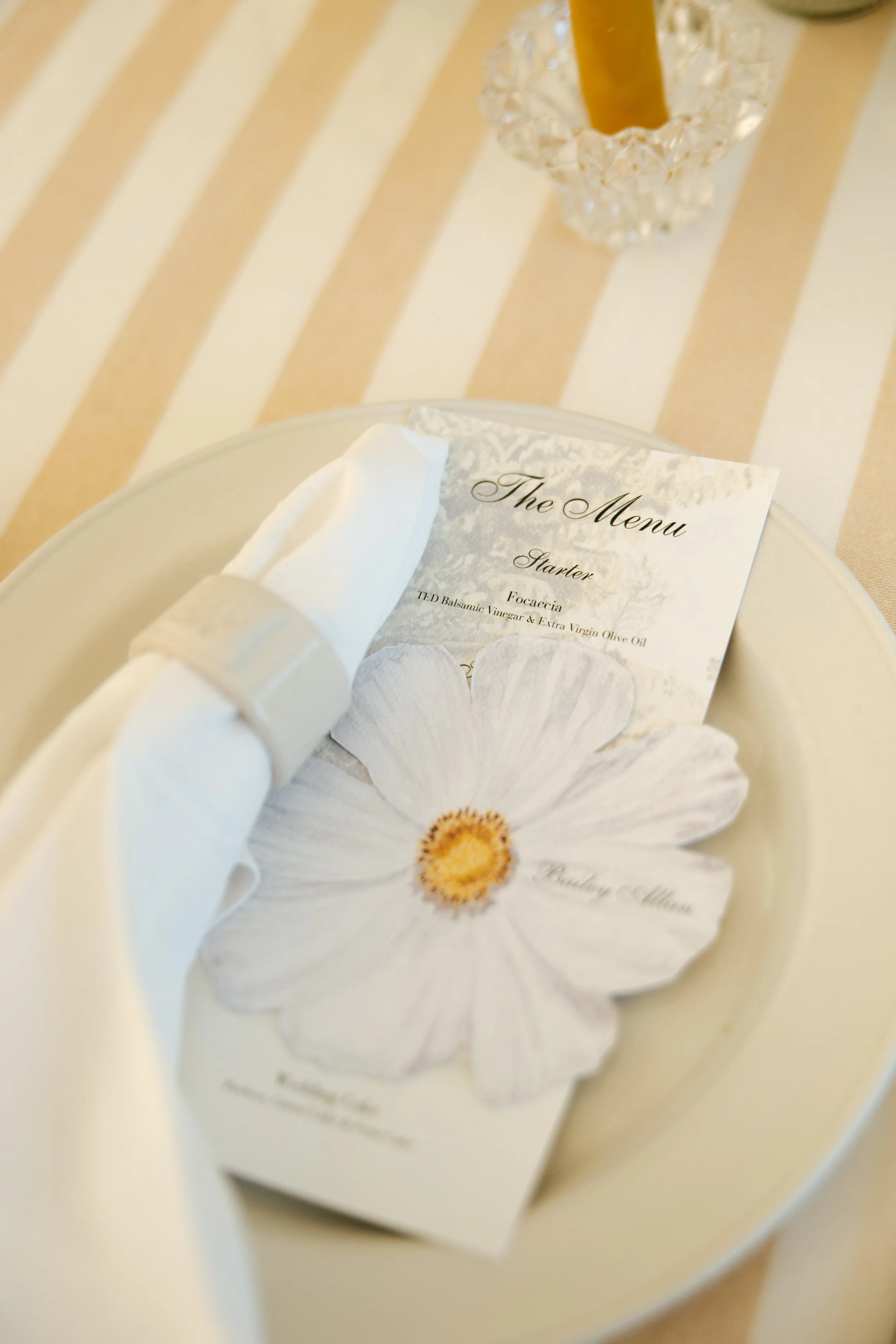 Close-up of a table setting with a cream-colored and beige striped tablecloth, a white napkin, a menu card, and a clear candle stick holder with a beeswax candlestick.