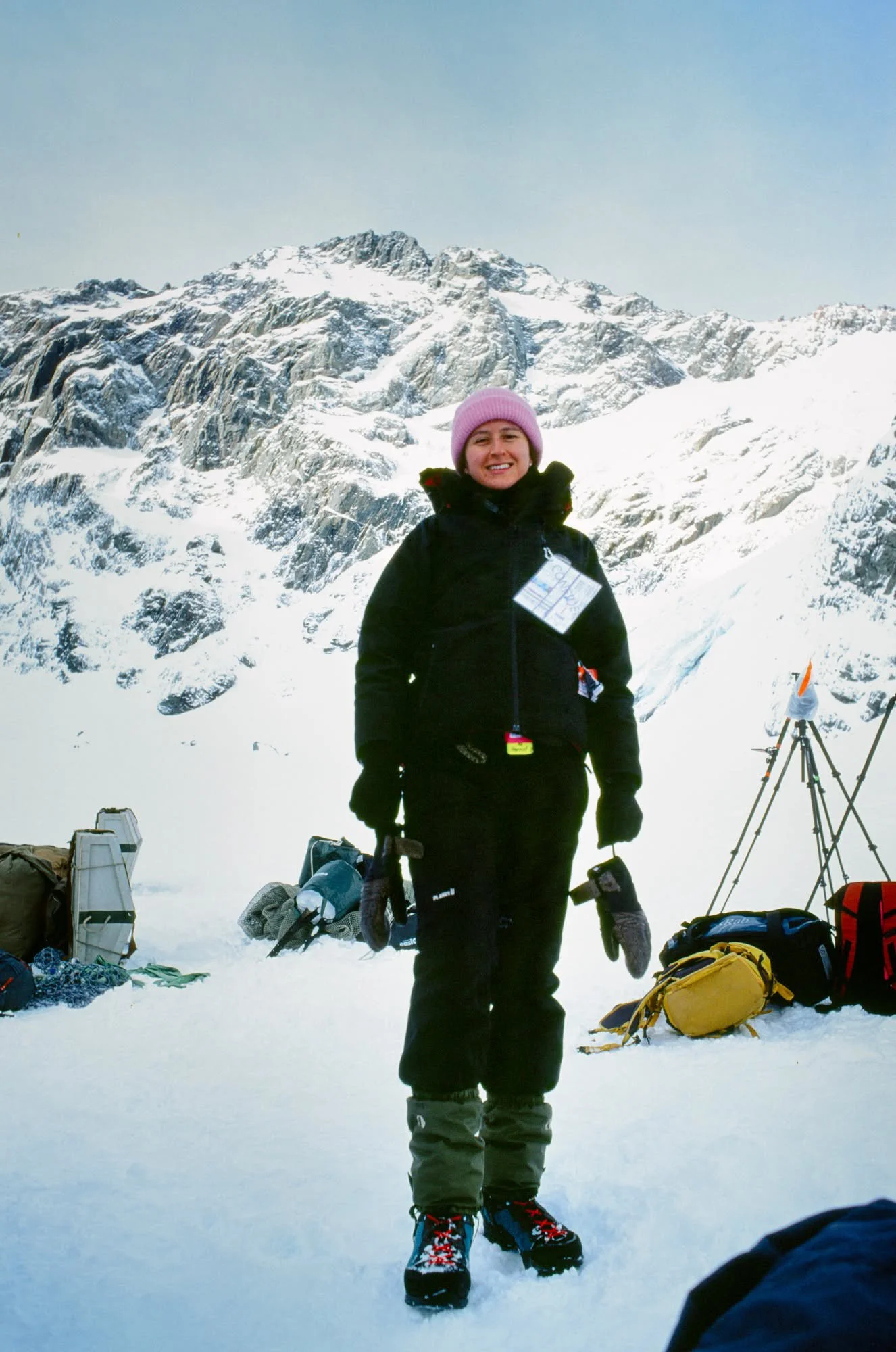 A woman standing in snow with mountain peaks in the background, wearing winter gear and holding gloves, surrounded by backpacks and equipment.