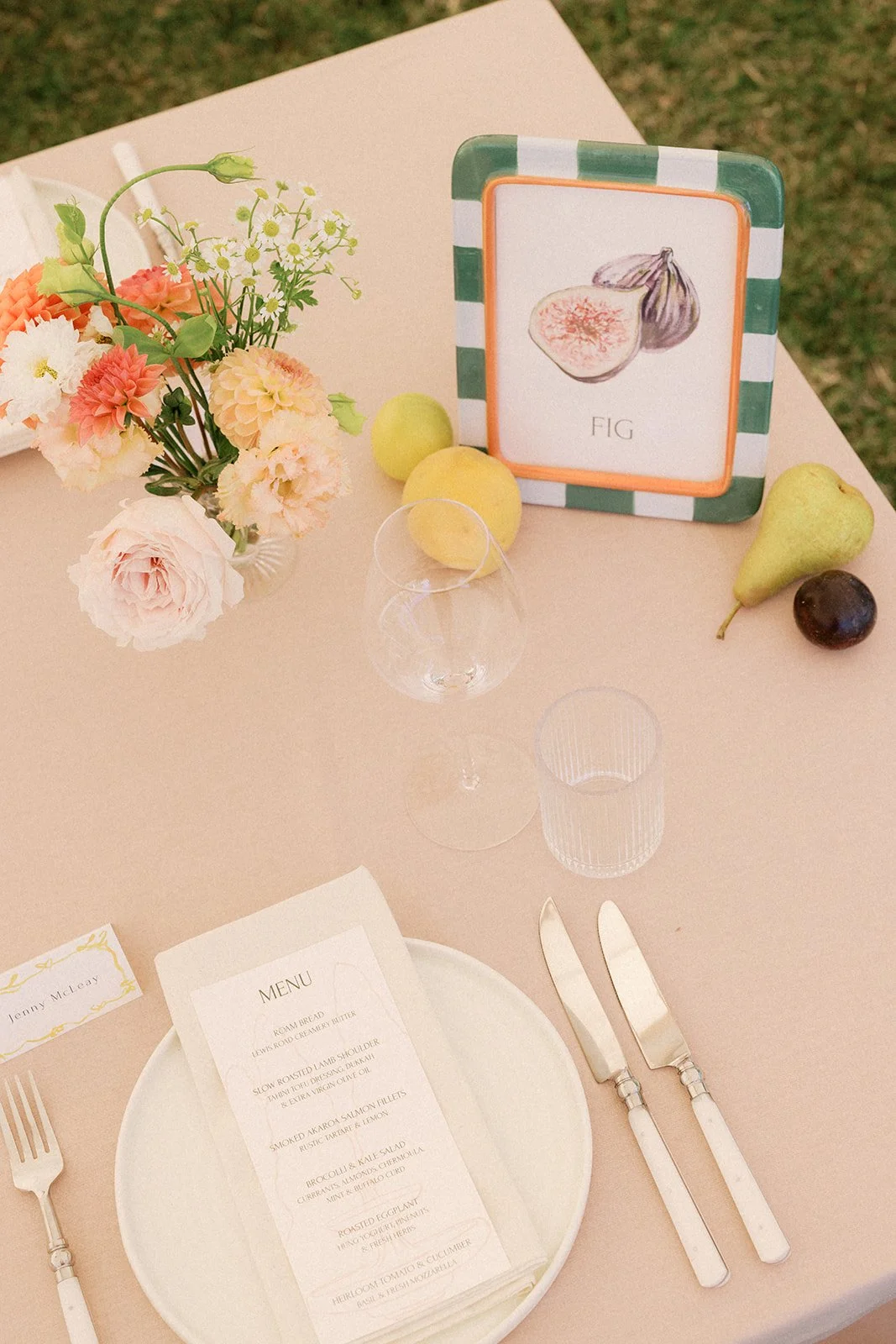 A table decorated for a meal with a pink tablecloth, a pink flower arrangement, an illustrated picture of a fig on a framed card, two pears, a plum, a glass, a knife and fork, and a menu on a white plate.